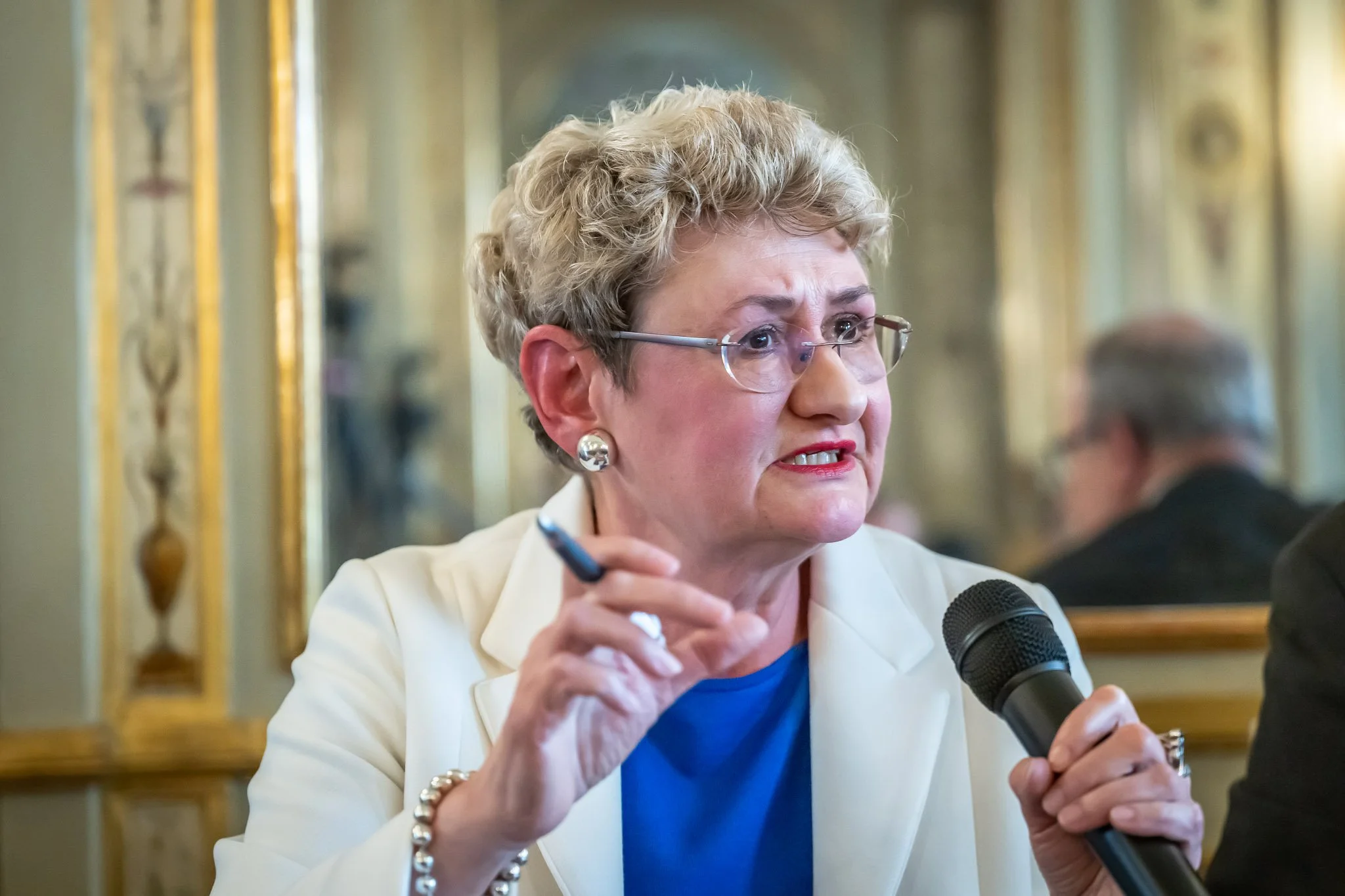 An elderly woman speaking into a microphone whilst holding a pen, wearing glasses and a white blazer, against a decorative background.