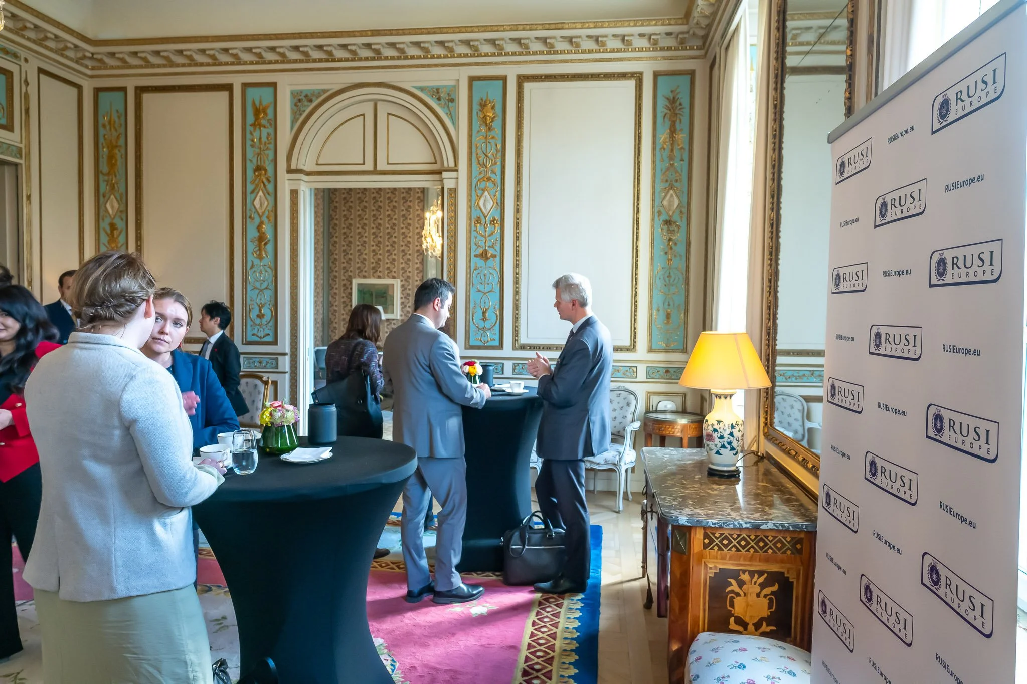 A group of people are standing and chatting in a decorated room at a networking event, with a ‘RUSI Europe’ banner visible on the right.