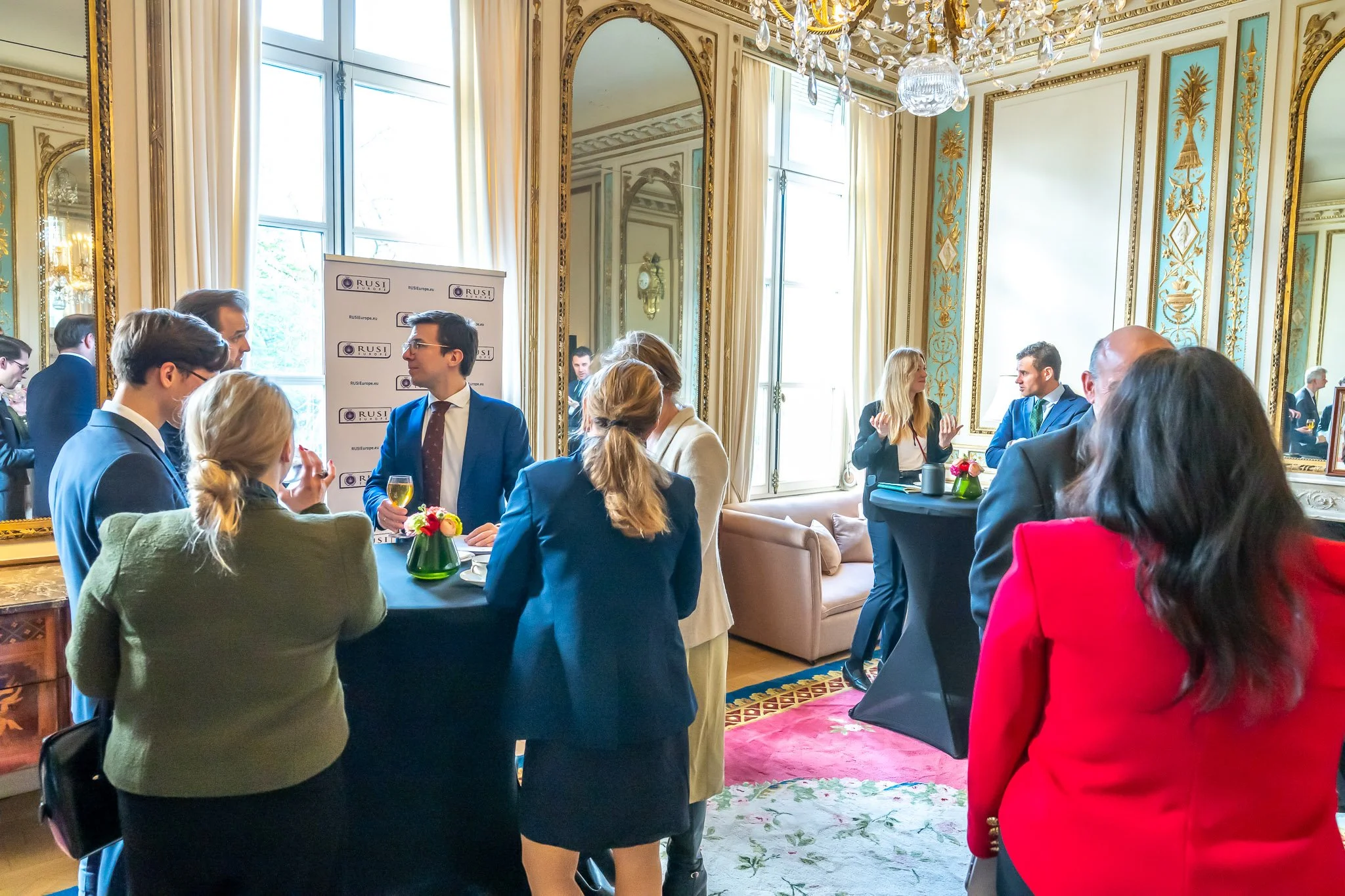 A group of people in formal attire are chatting in an elegant lounge with mirrors, a chandelier and curtains. Drinks and floral arrangements are arranged on small, tall tables."}