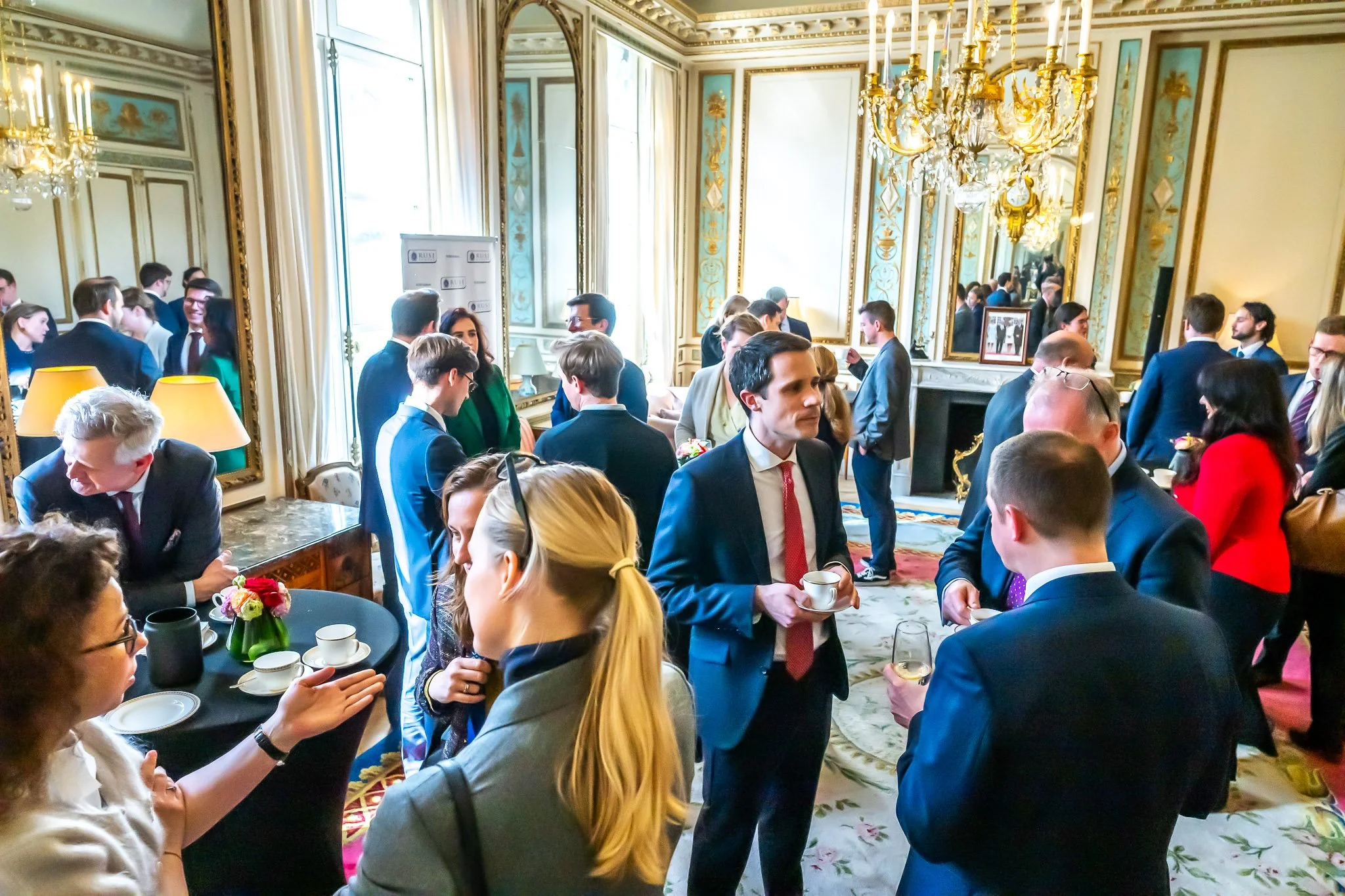 Conference participants converse over refreshments in an ornate room with mirrors and chandeliers.