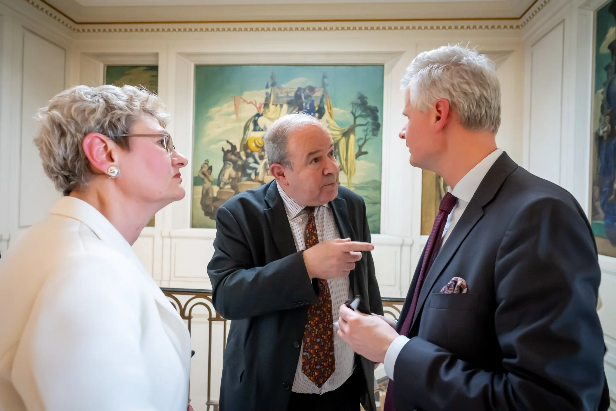 Three people in suits, engaged in a lively discussion in a room decorated with large murals.