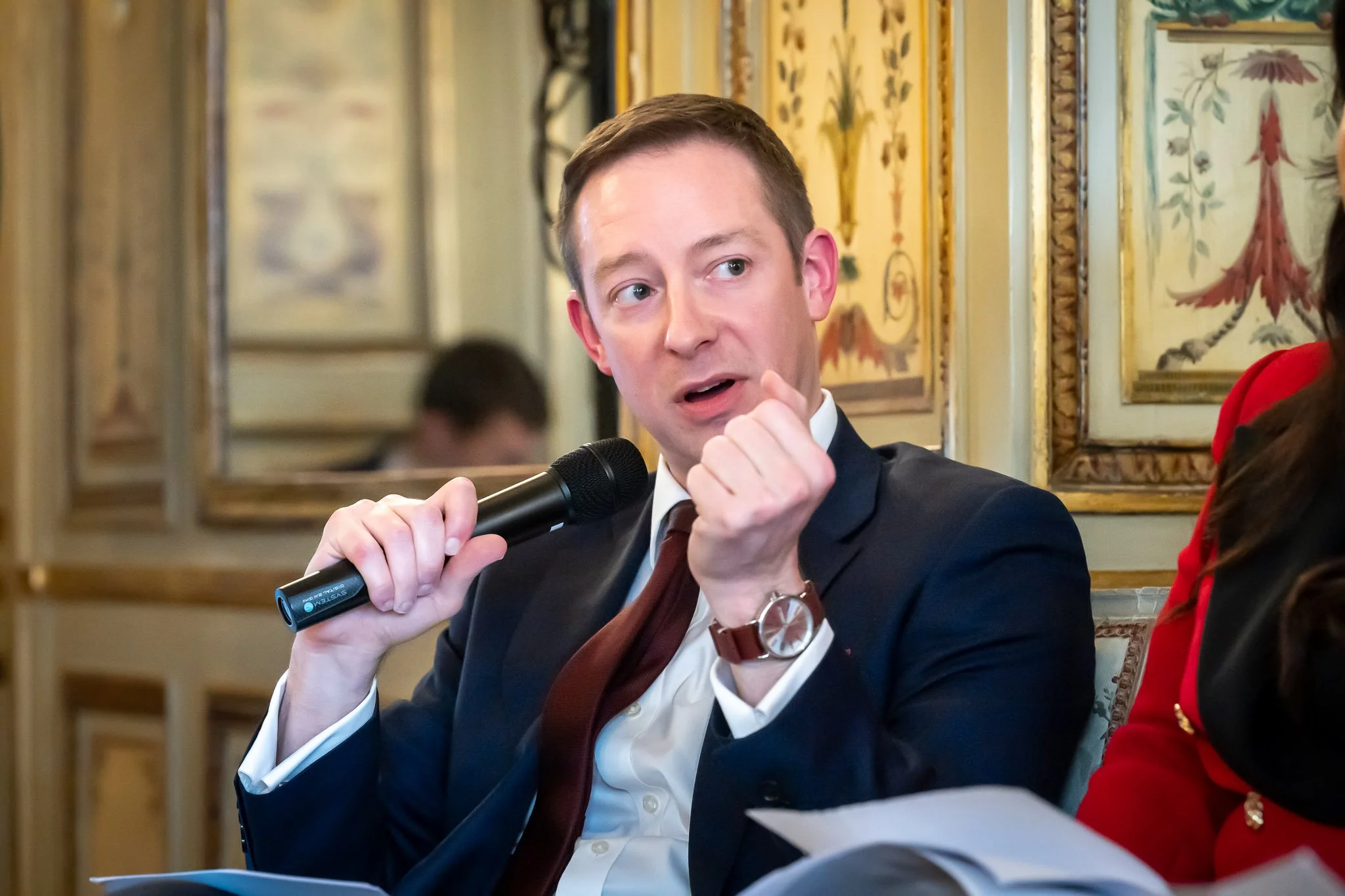 A man in a suit, holding a microphone, is taking part in a discussion in an ornate room.