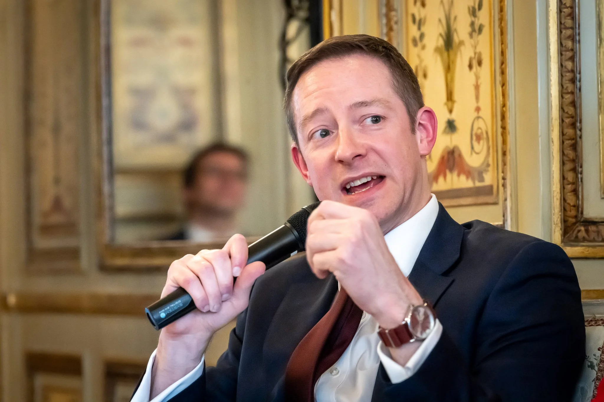 A man in a suit holding a microphone, speaking at an event or conference, with an elegant interior setting in the background.