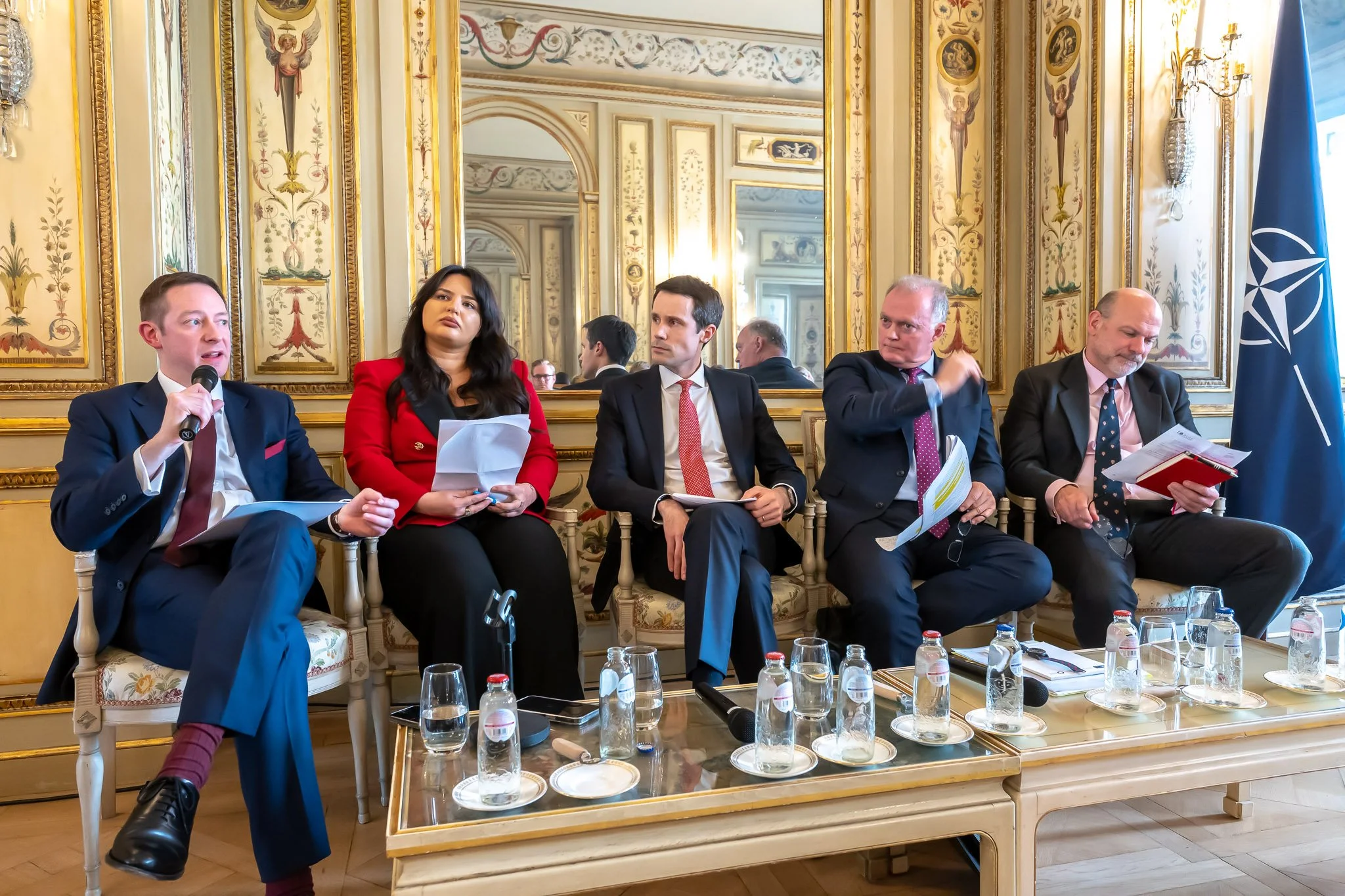Five people are sitting in an elegant lounge, chatting, with documents and bottles of water on a coffee table. A NATO flag is visible on the right. The décor is richly ornate, with a mirror in the background.