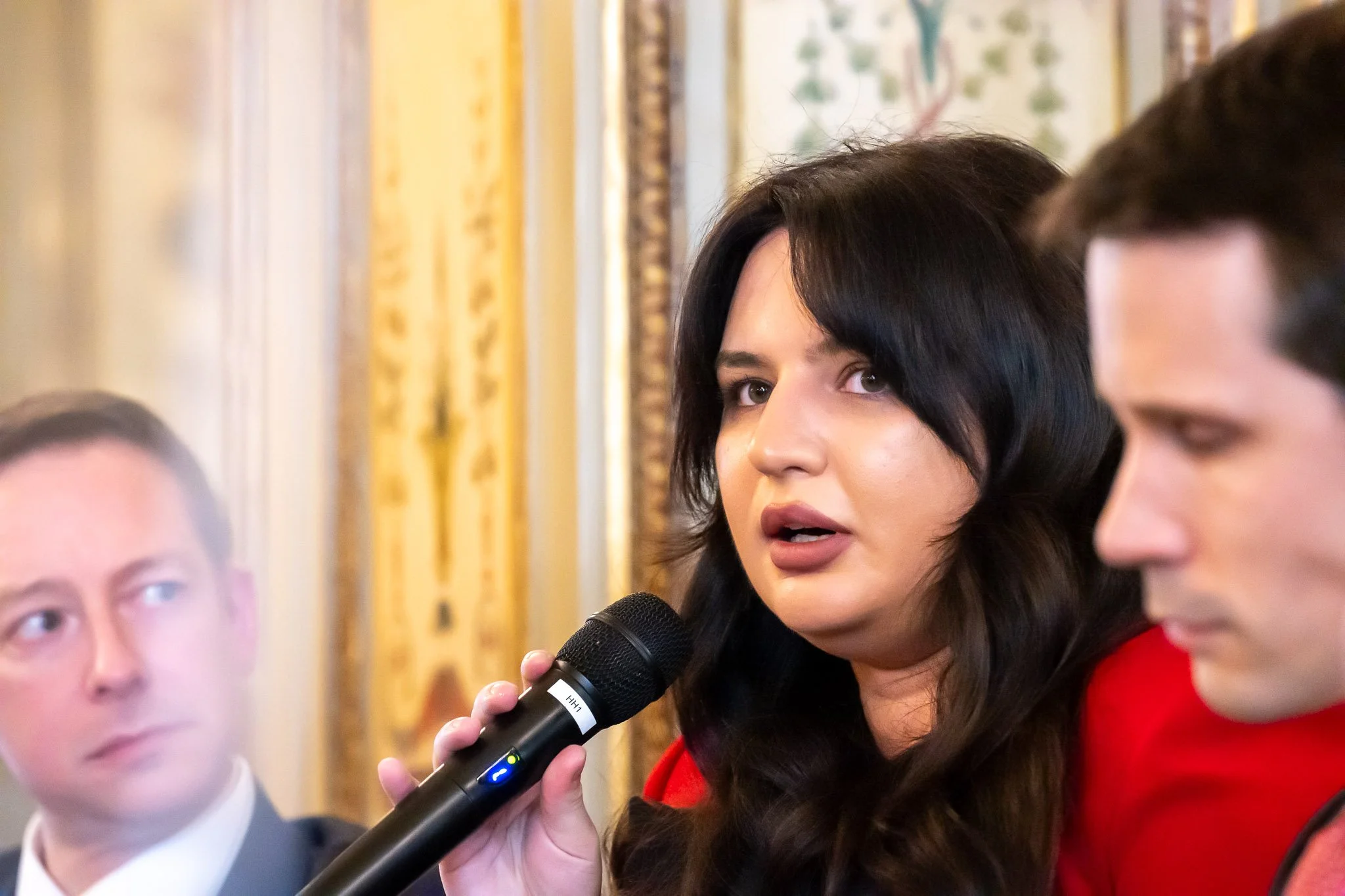 A person speaking into a microphone, looking serious, with two other people listening attentively in the background, against an elegant interior setting.