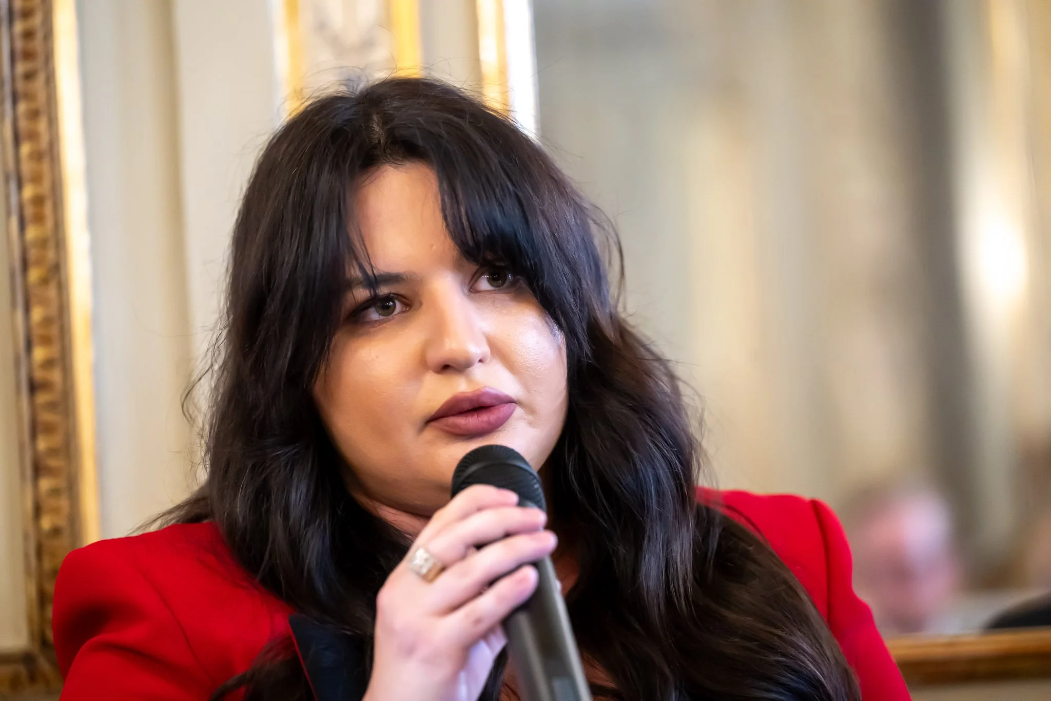 A person speaking into a microphone, with a blurred background, wearing a red blazer.