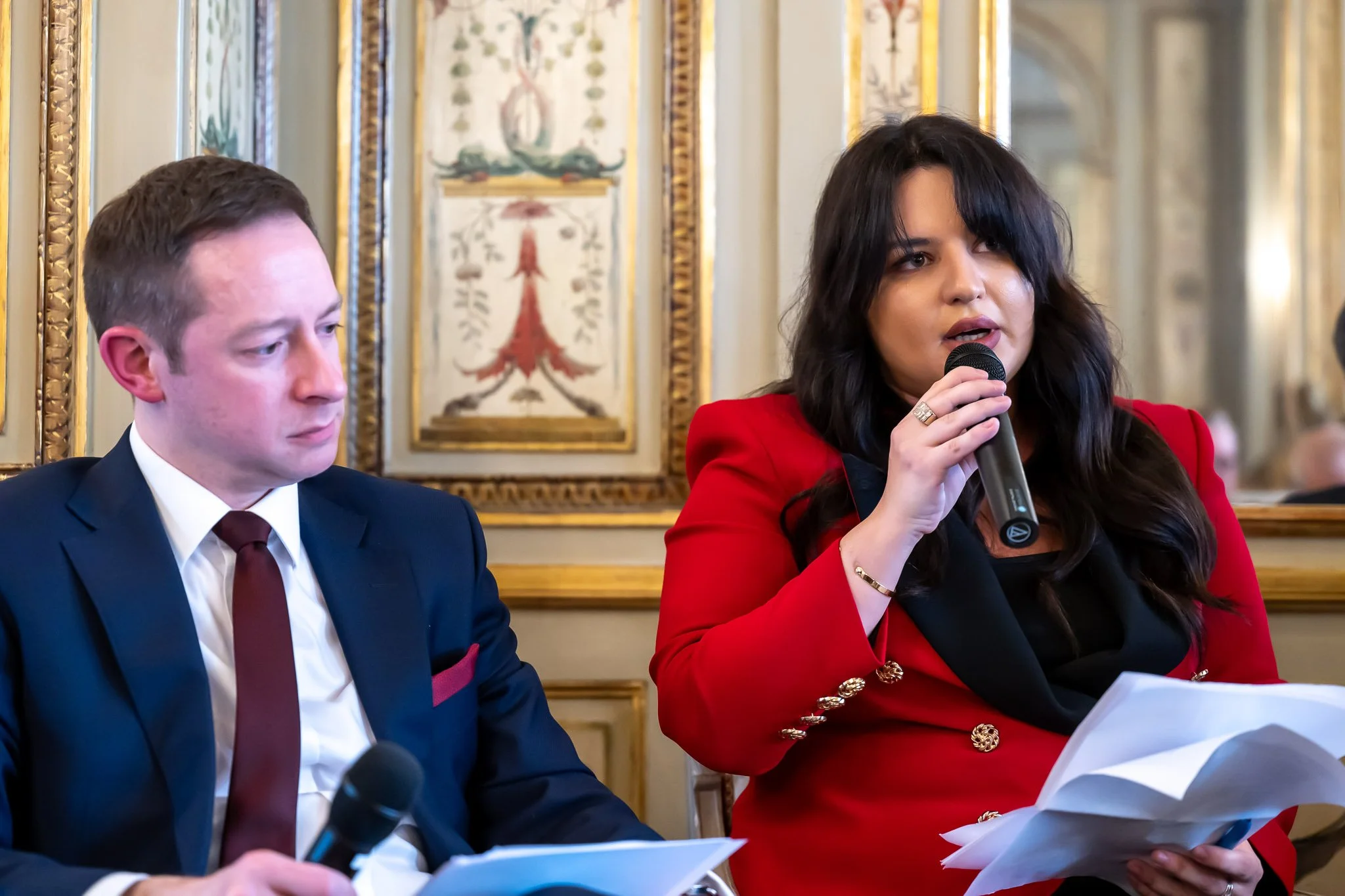 A man in a suit sitting next to a woman in a red jacket, holding a microphone and some papers, in a room decorated with classical motifs.
