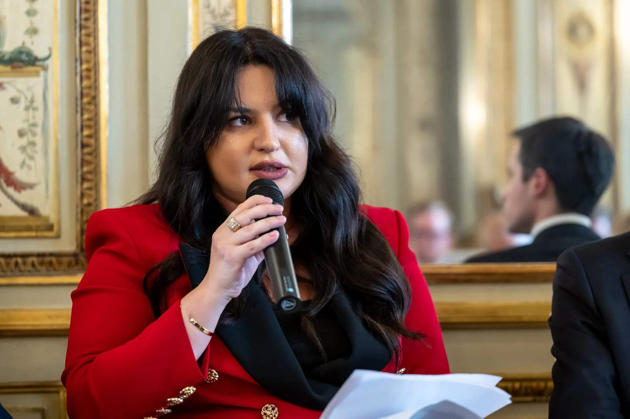 A woman in a red jacket, holding a microphone, sitting in an ornately decorated room.