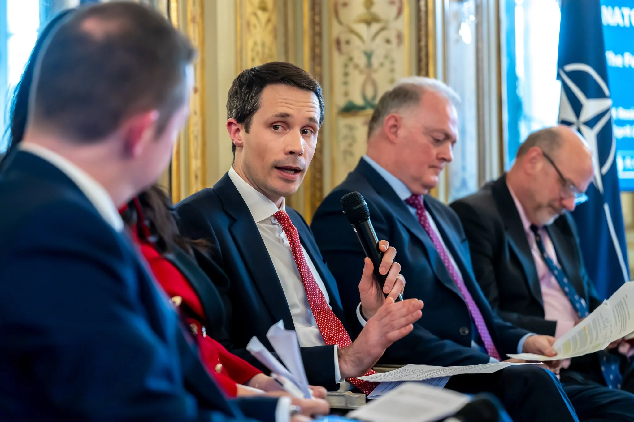 A group of people in formal attire are taking part in a discussion or a conference. One of them is holding a microphone and speaking, whilst the others are listening or taking notes. A NATO flag can be seen in the background.