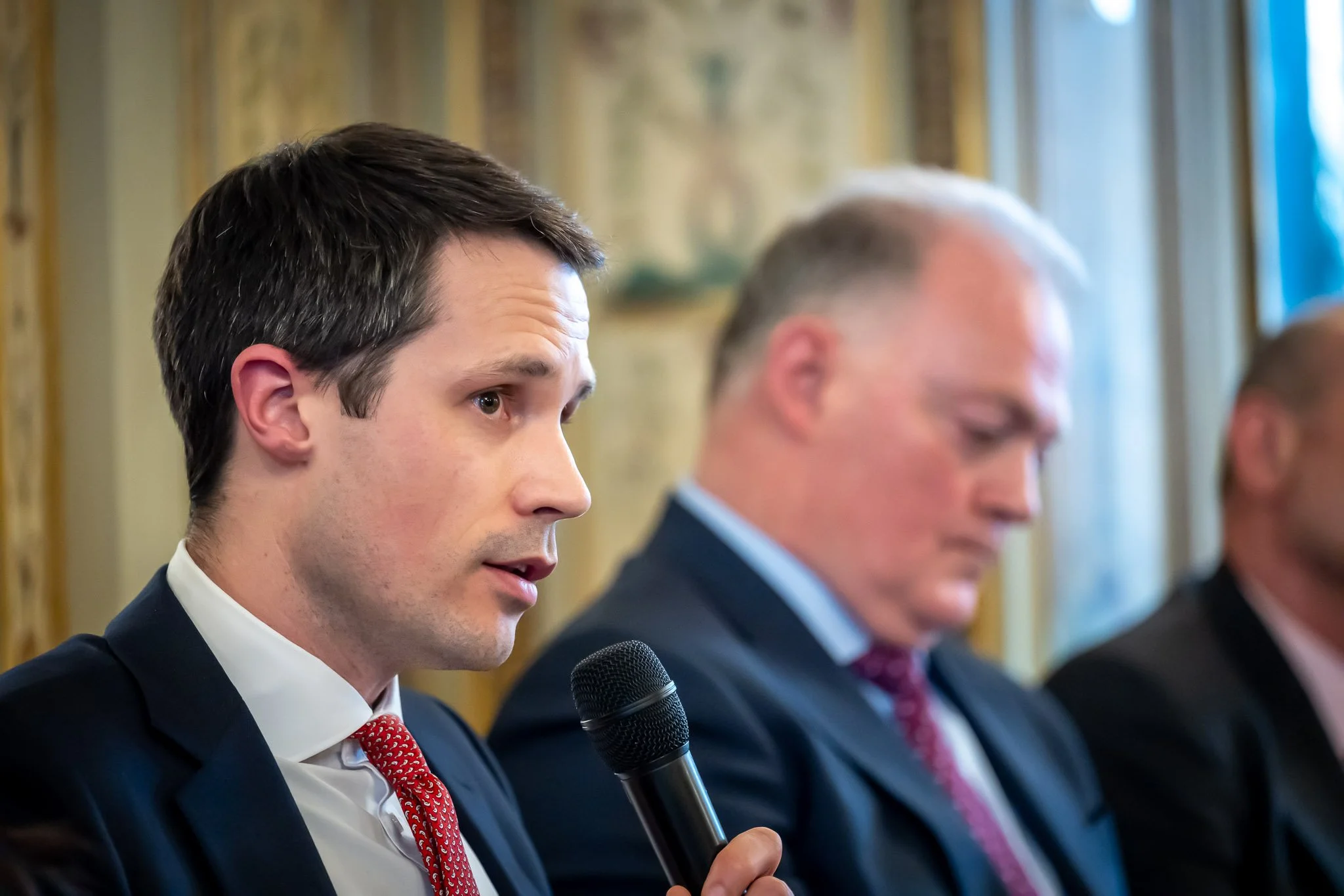 A man in a suit is speaking into a microphone at a conference, with another man in a suit in the background.
