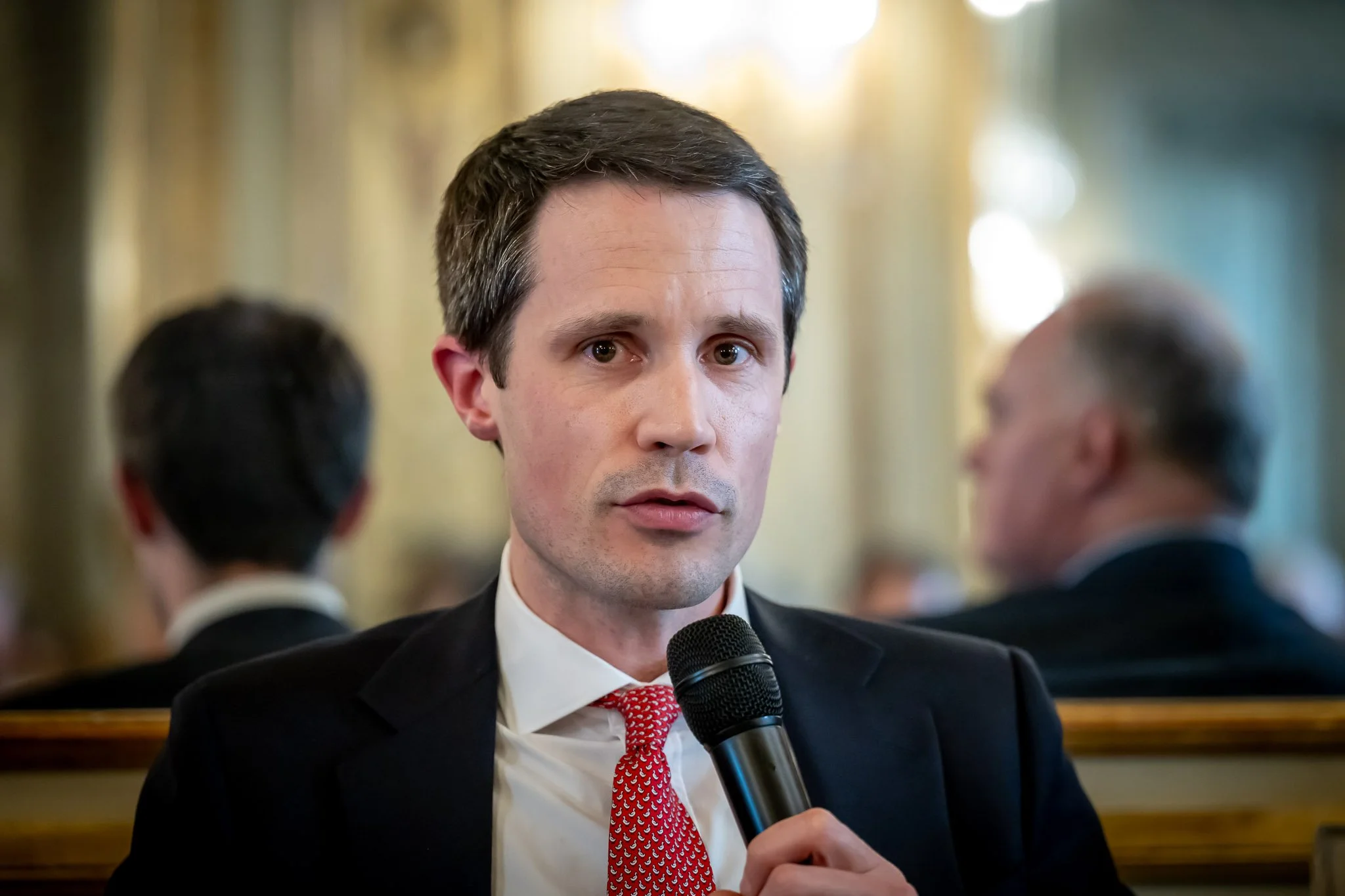 A man in a suit holding a microphone, speaking at an event.