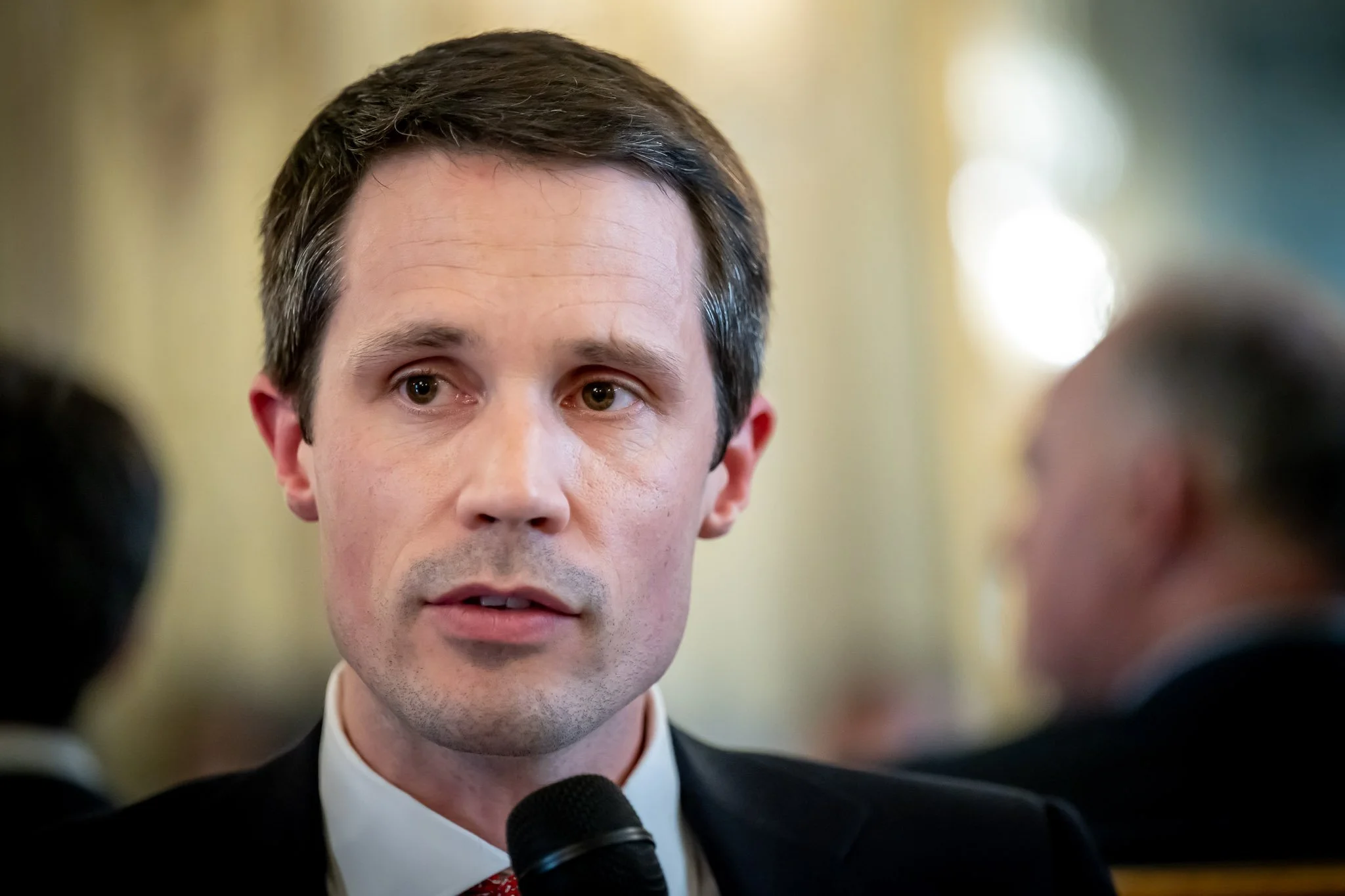 A man in a suit is speaking into a microphone, against a blurred background.