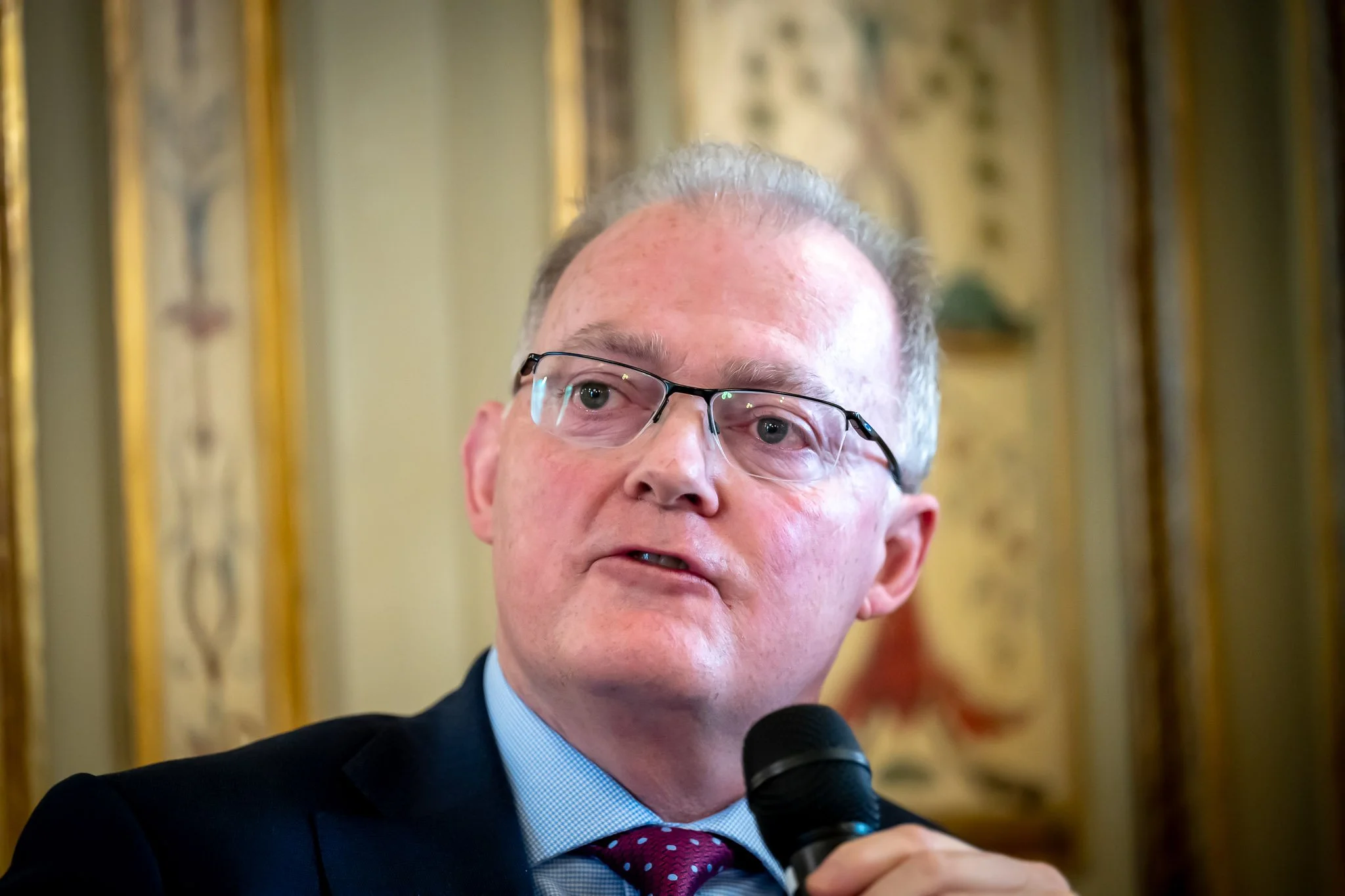 A man wearing glasses speaking into a microphone, against a decorative backdrop.