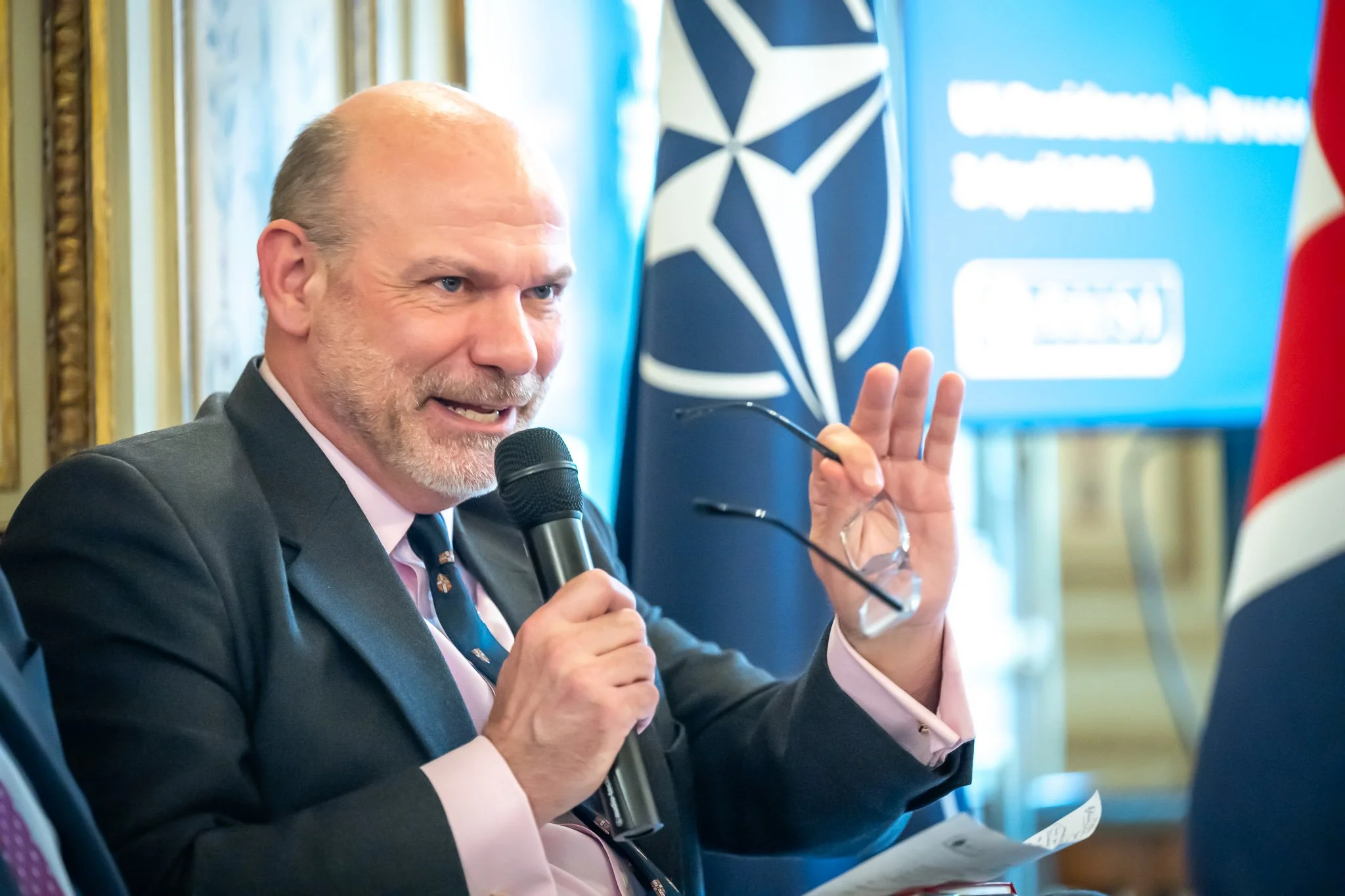 A man holding a microphone and speaking at an event, with a NATO flag and logo in the background.