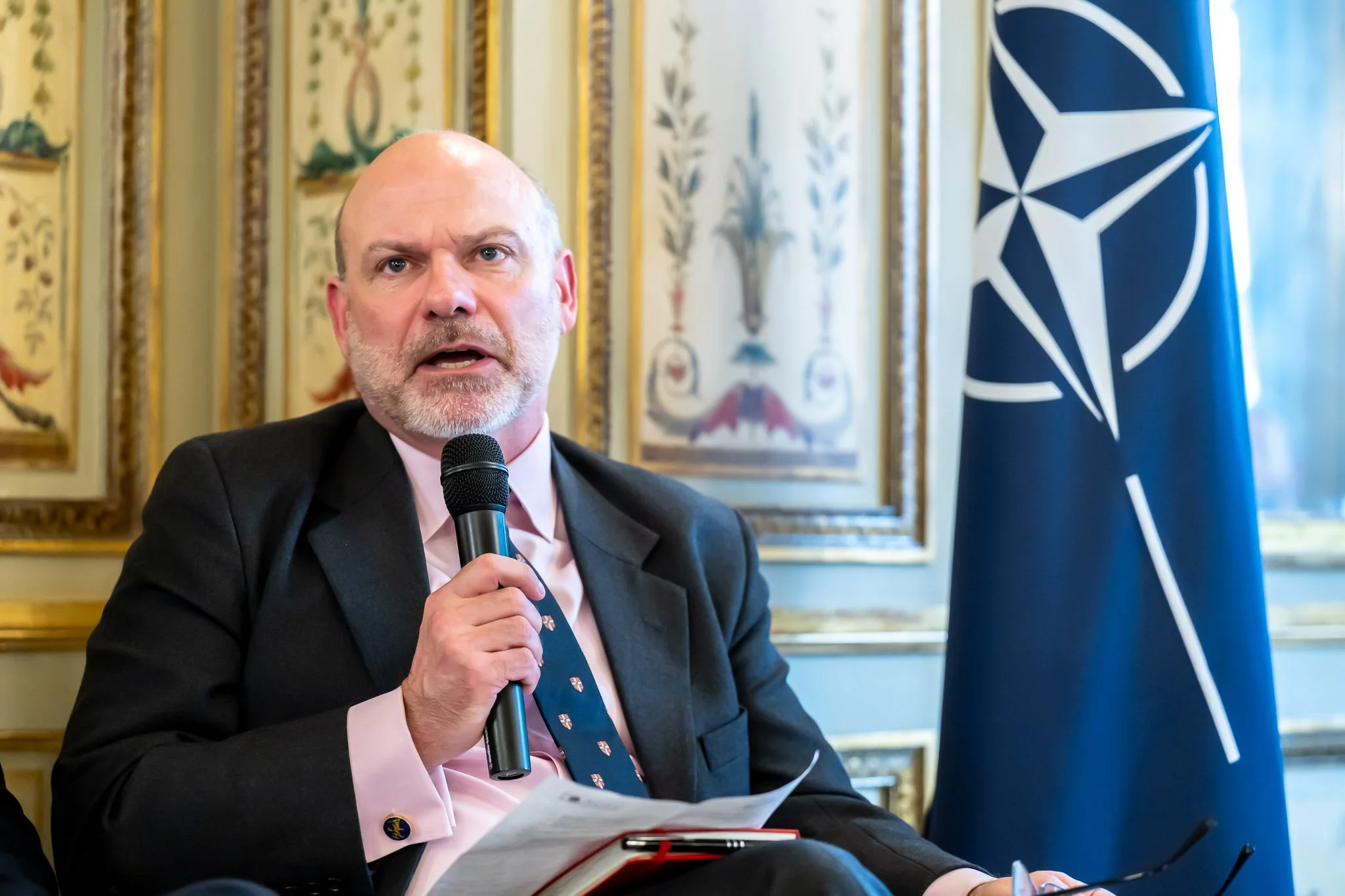 A man in a suit speaking into a microphone, with a NATO flag next to him.