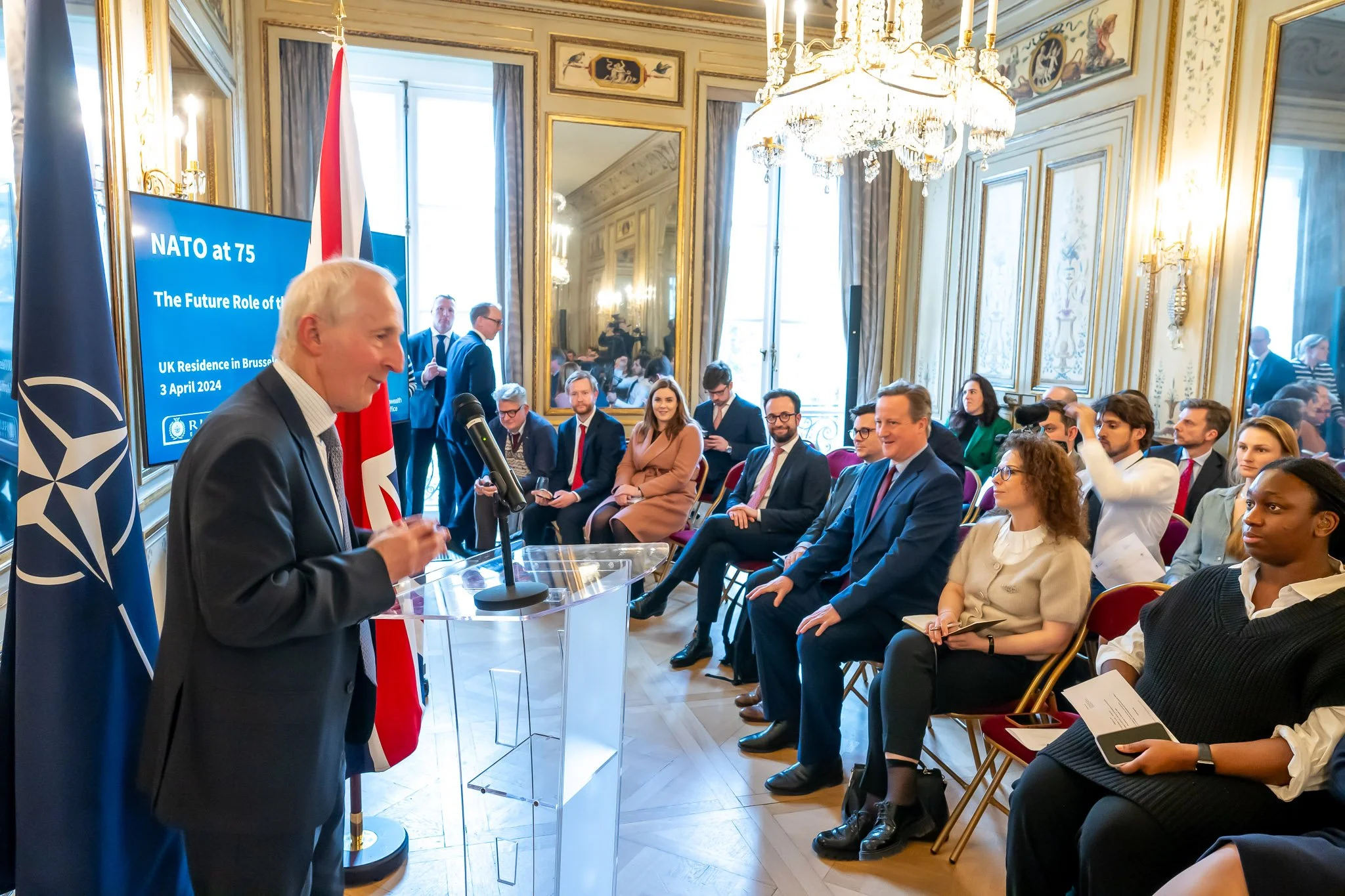 A conference is taking place in an elegant room with a speaker at the microphone. A screen displays ‘NATO at 75’ and ‘The Future Role of the UK’. The participants are seated and listening attentively. Flags and mirrors adorn the room.