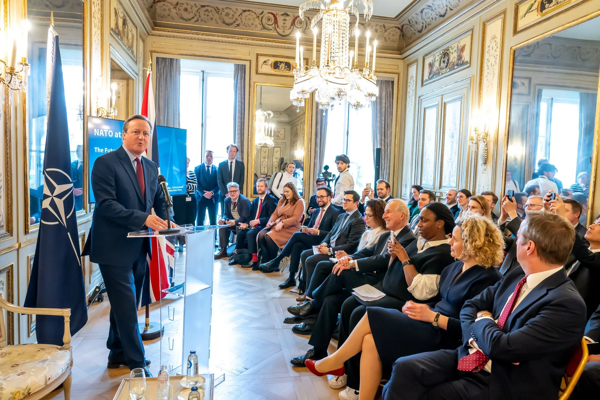 A man in a suit speaking to an audience in a room decorated with the NATO flag. The seated audience is listening attentively; some are taking photographs. Candlesticks and decorative mirrors can be seen in the background.
