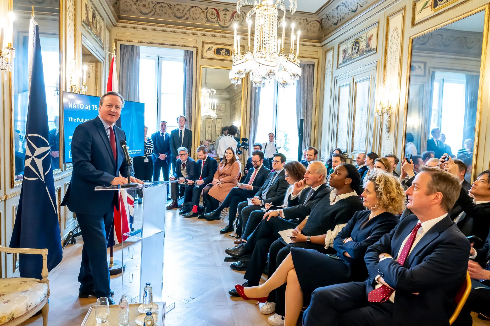 A man in a suit is giving a speech to an audience seated in an elegant hall decorated with flags and the NATO logo. A presentation on the screen discusses the future of security.