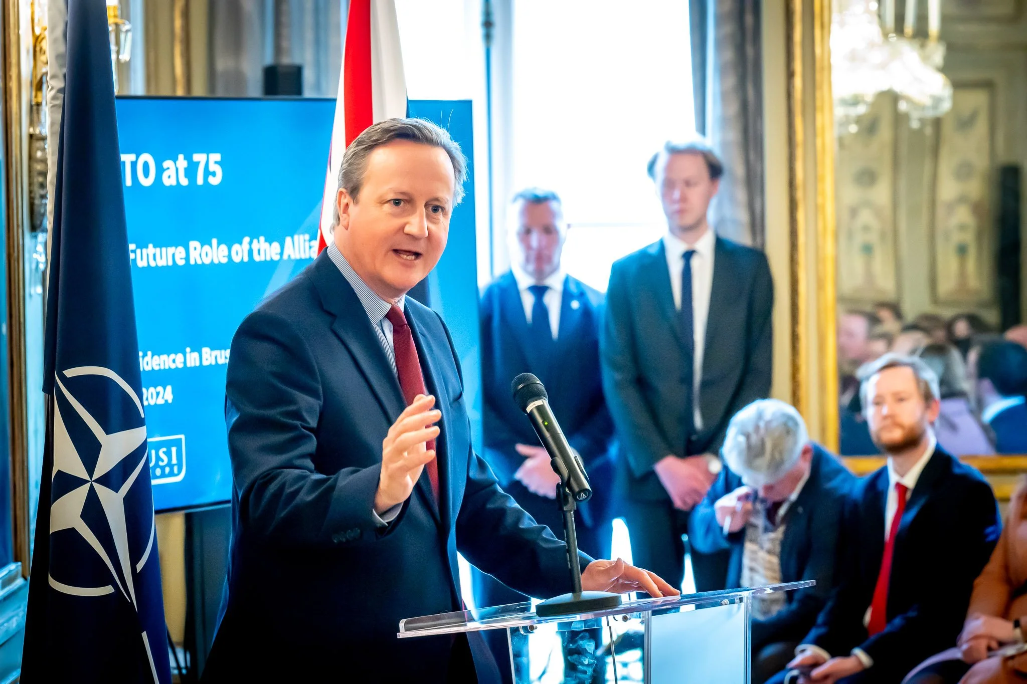 A man in a suit speaking into a microphone at a NATO-related event, with a screen in the background displaying ‘NATO at 75’. Several people in suits are seated and standing in the background, with a NATO flag visible on the left.