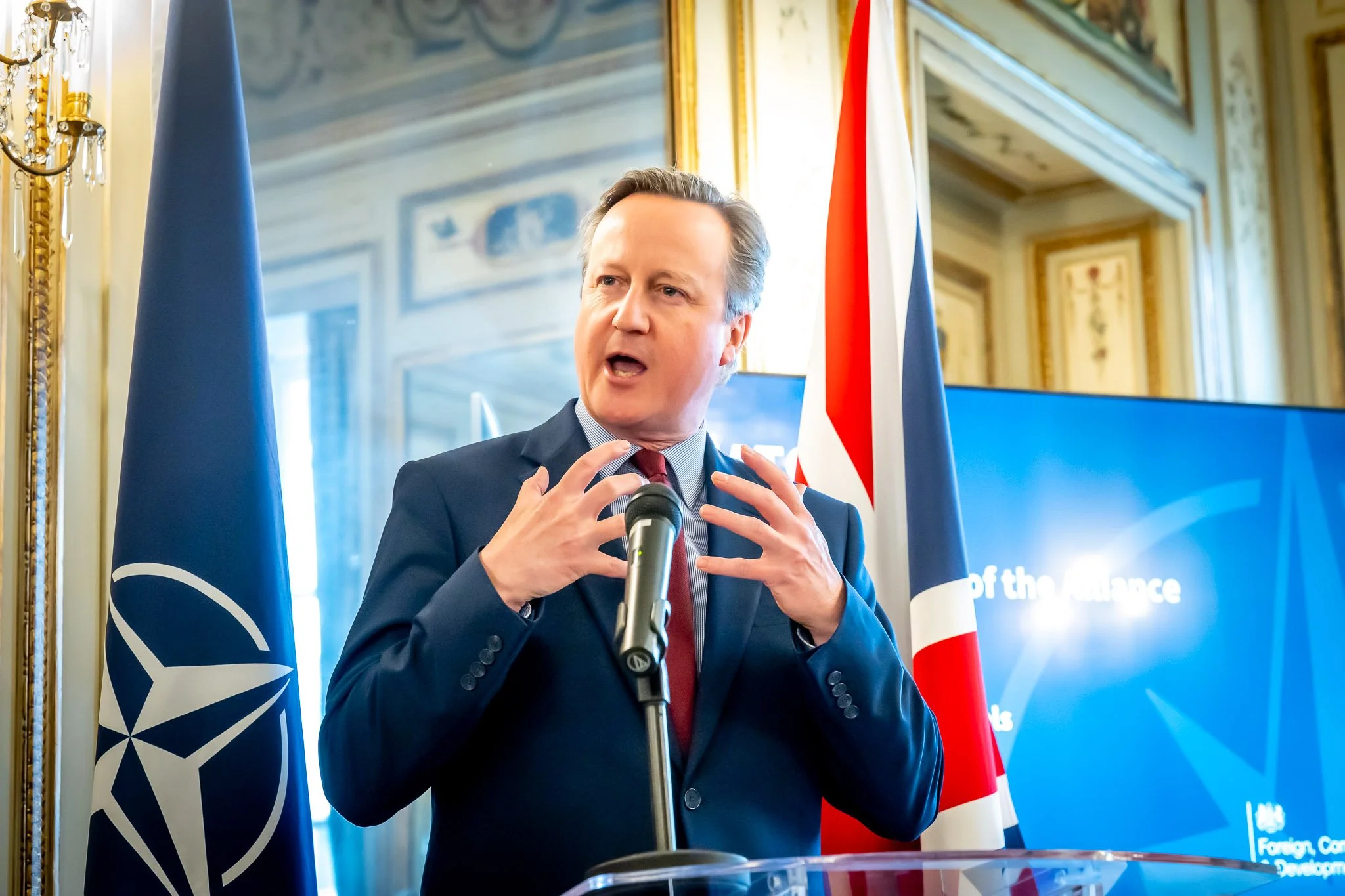 A man in a suit is giving a speech at a microphone, with a Union Jack and a NATO flag beside him. He is standing in an ornately decorated room.