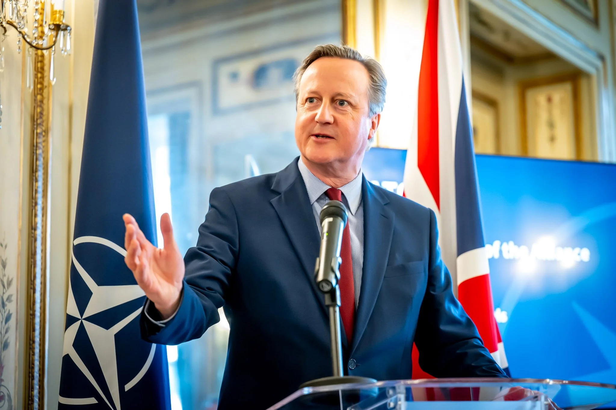Photo professionnelle de David Cameron devant un micro avec drapeaux de l'OTAN et du Royaume-Uni en arrière-plan.