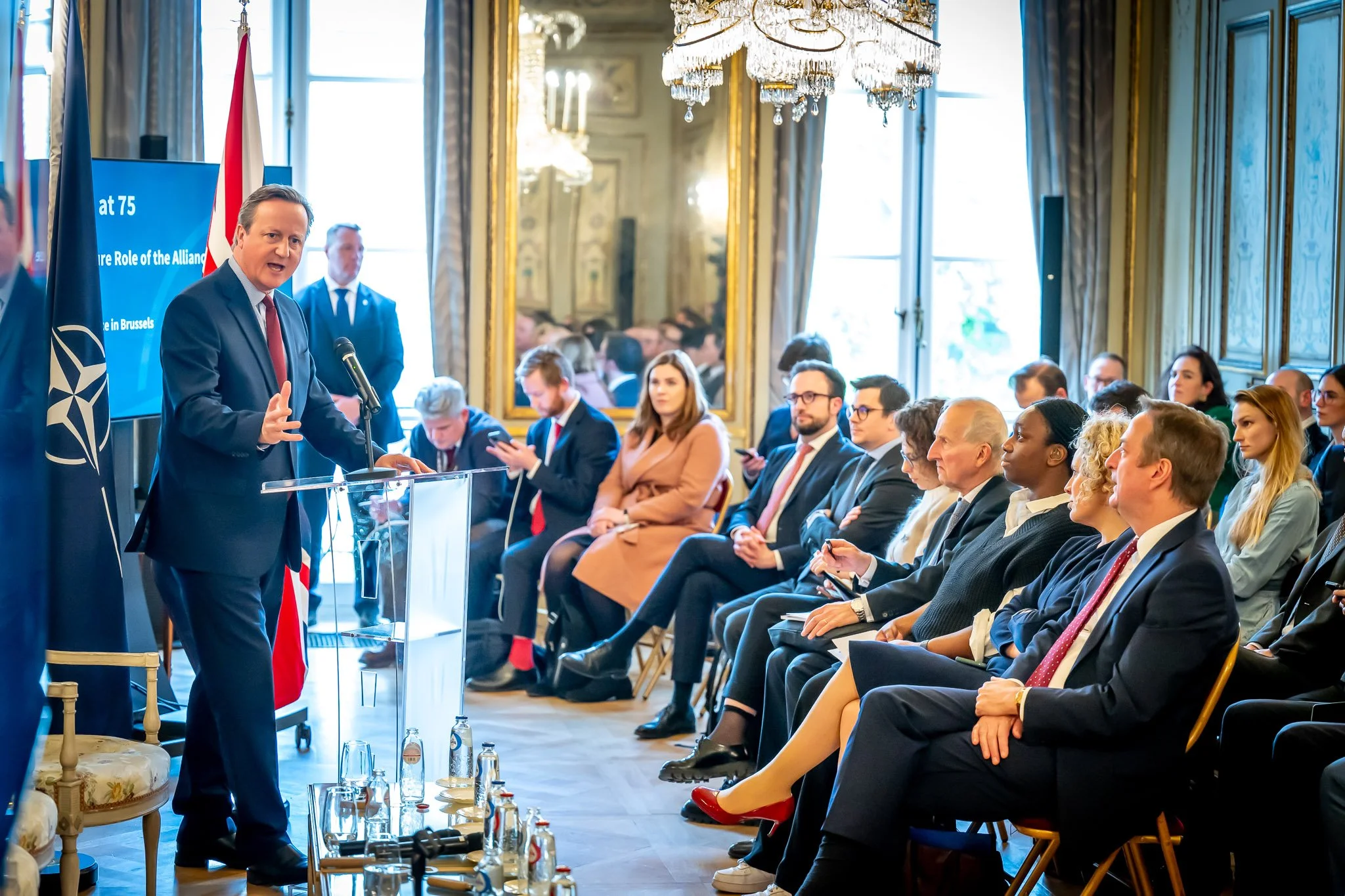 A man in a suit is speaking to an audience at a conference, with a NATO flag and a screen in the background. The attendees are listening attentively and are seated in an elegant room with a chandelier.