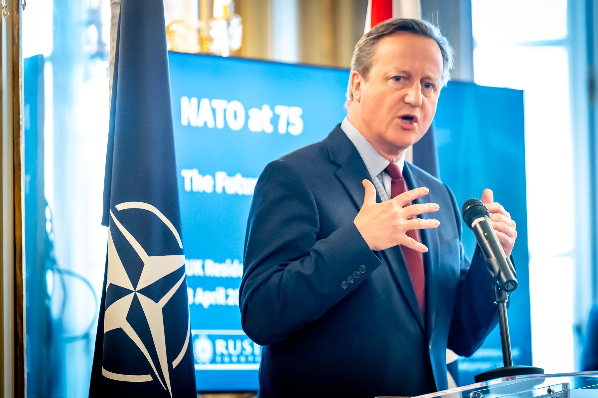 A man in a suit is speaking into a microphone at an event marking NATO’s 75th anniversary, with a NATO flag beside him.