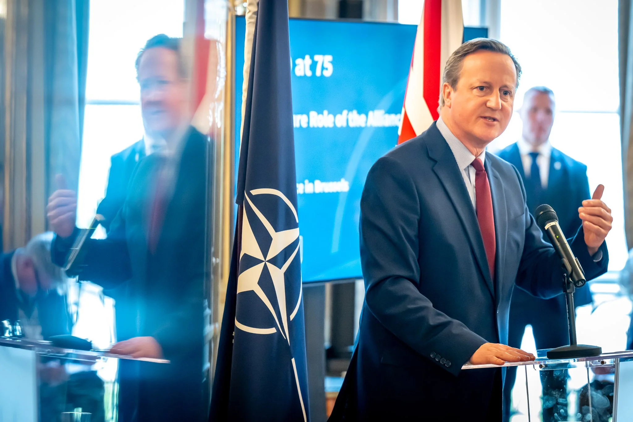 A man in a suit is speaking into a microphone in front of a NATO flag at a conference or official event. A screen in the background displays information relating to NATO.