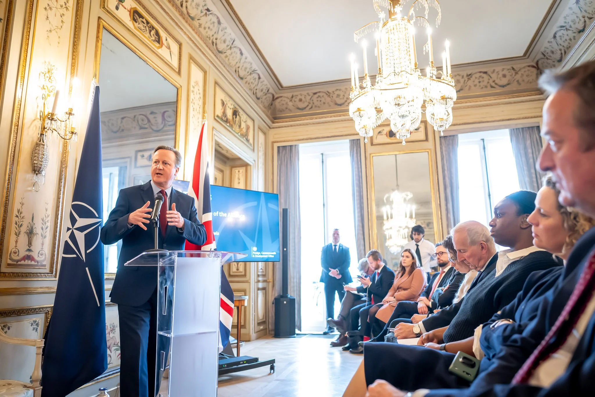 A man in a suit speaking from a podium at a conference in an elegant room featuring a chandelier, the Union Jack and the NATO flag, with an audience seated and listening.