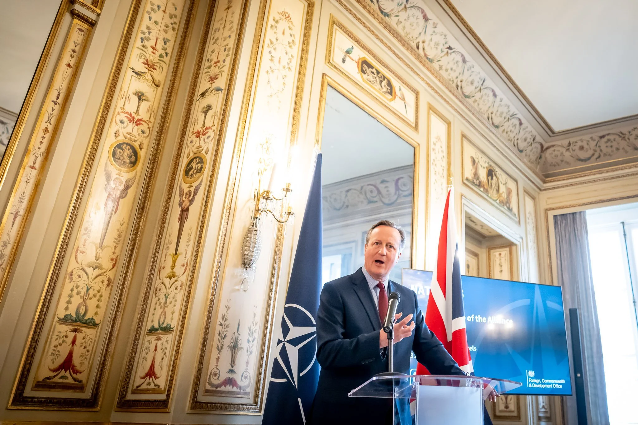 A man in a suit speaking from a lectern, with NATO and UK flags in the background, inside a decorated room.