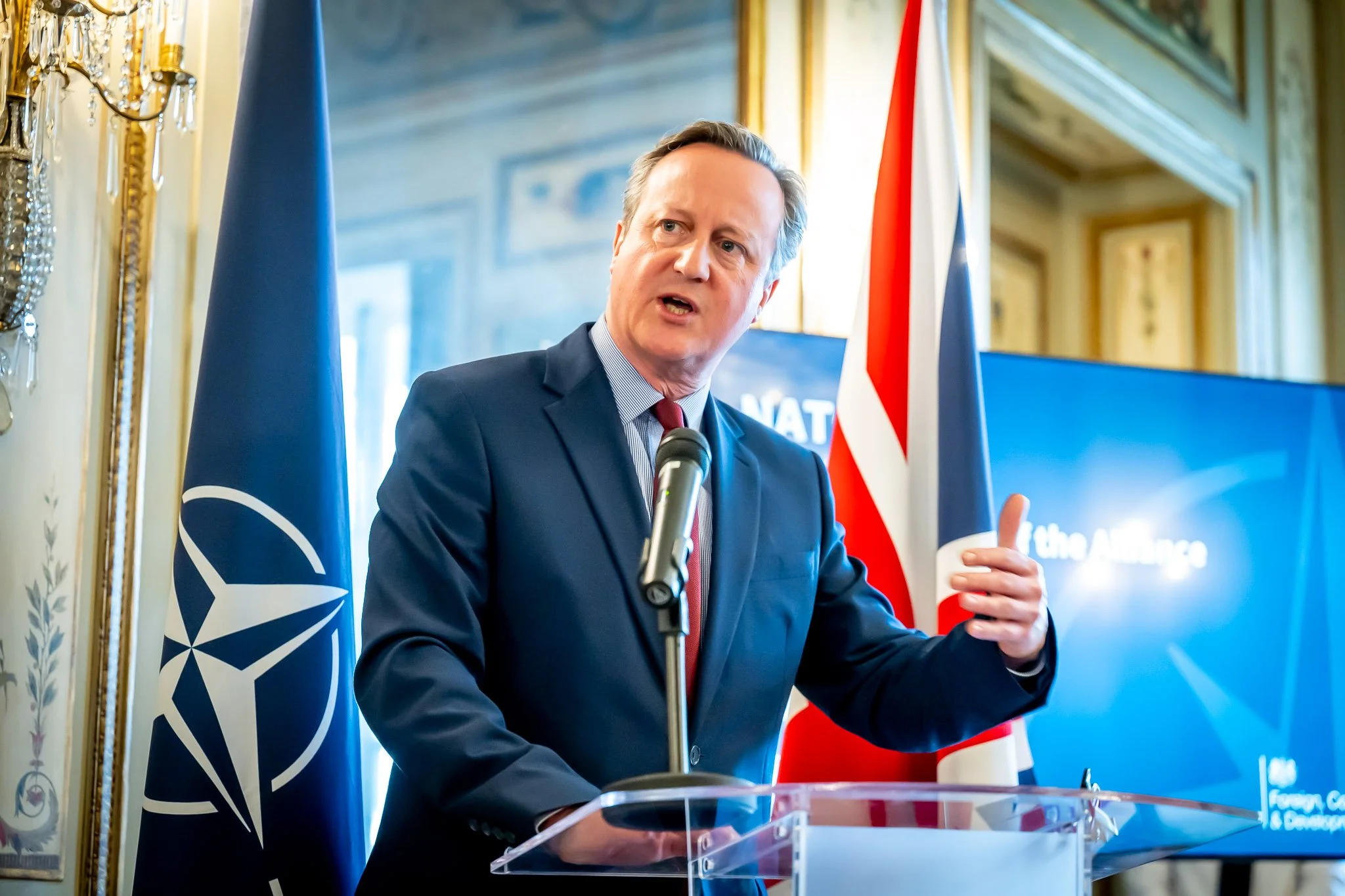 A man in a suit giving a speech in front of NATO and UK flags.