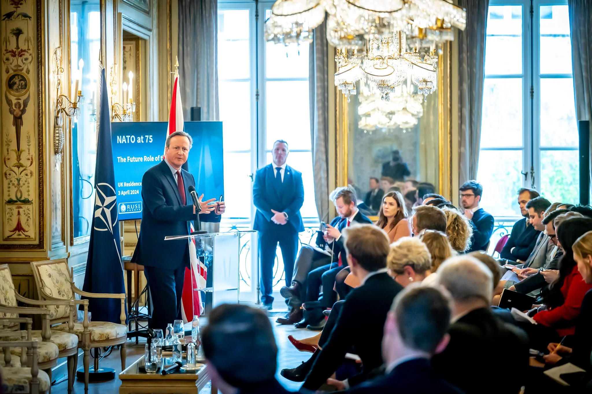 A conference marking NATO’s 75th anniversary, featuring a speaker addressing an audience in a hall adorned with chandeliers. A screen displays the words ‘NATO at 75’.