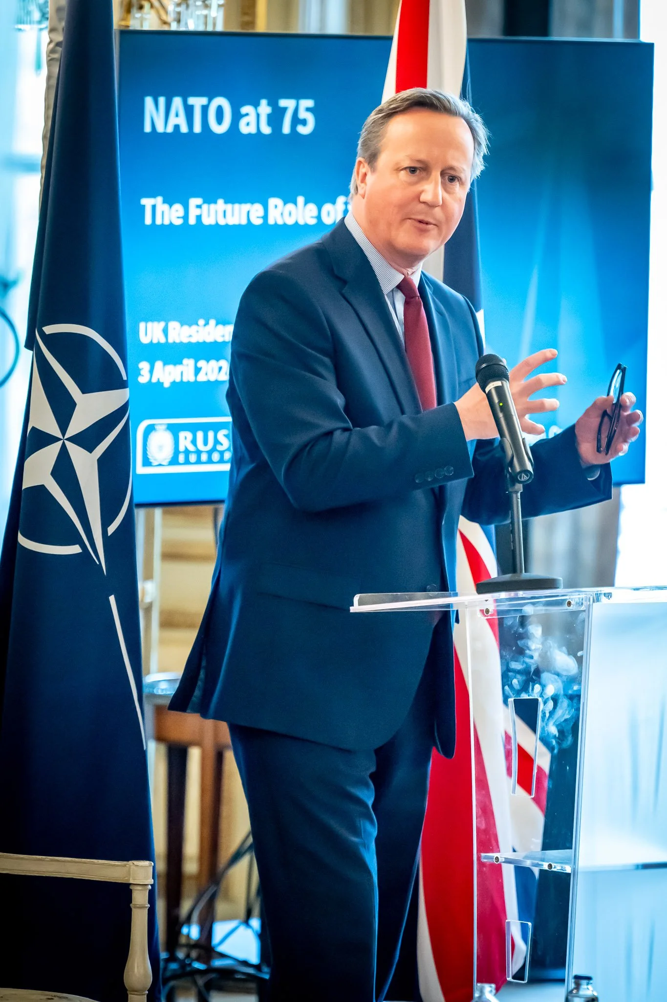 A man in a suit is speaking at a lectern with a screen and a flag behind him. The screen displays ‘NATO at 75’. A microphone is attached to the lectern and a NATO flag can be seen next to it.