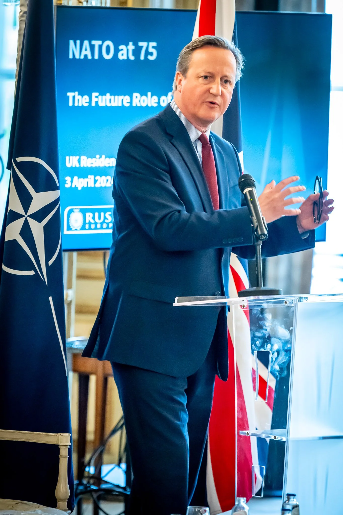 Photographie lors d'un événement institutionnel. David Cameron en costume donne une présentation près d'un drapeau de l'OTAN et d'un drapeau national. En arrière-plan, on voit un écran avec le texte "NATO at 75".