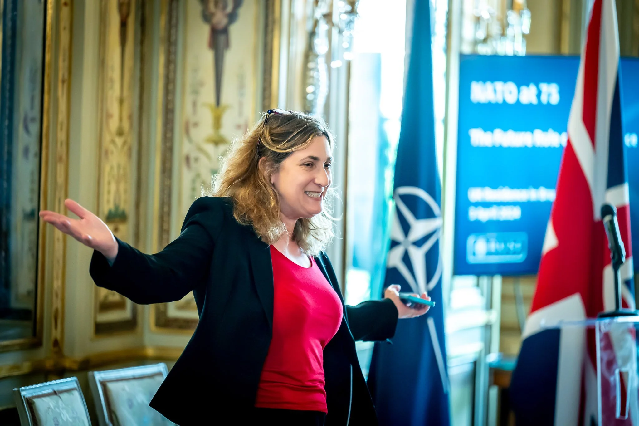 A woman presenting at an event, with NATO and UK flags in the background.