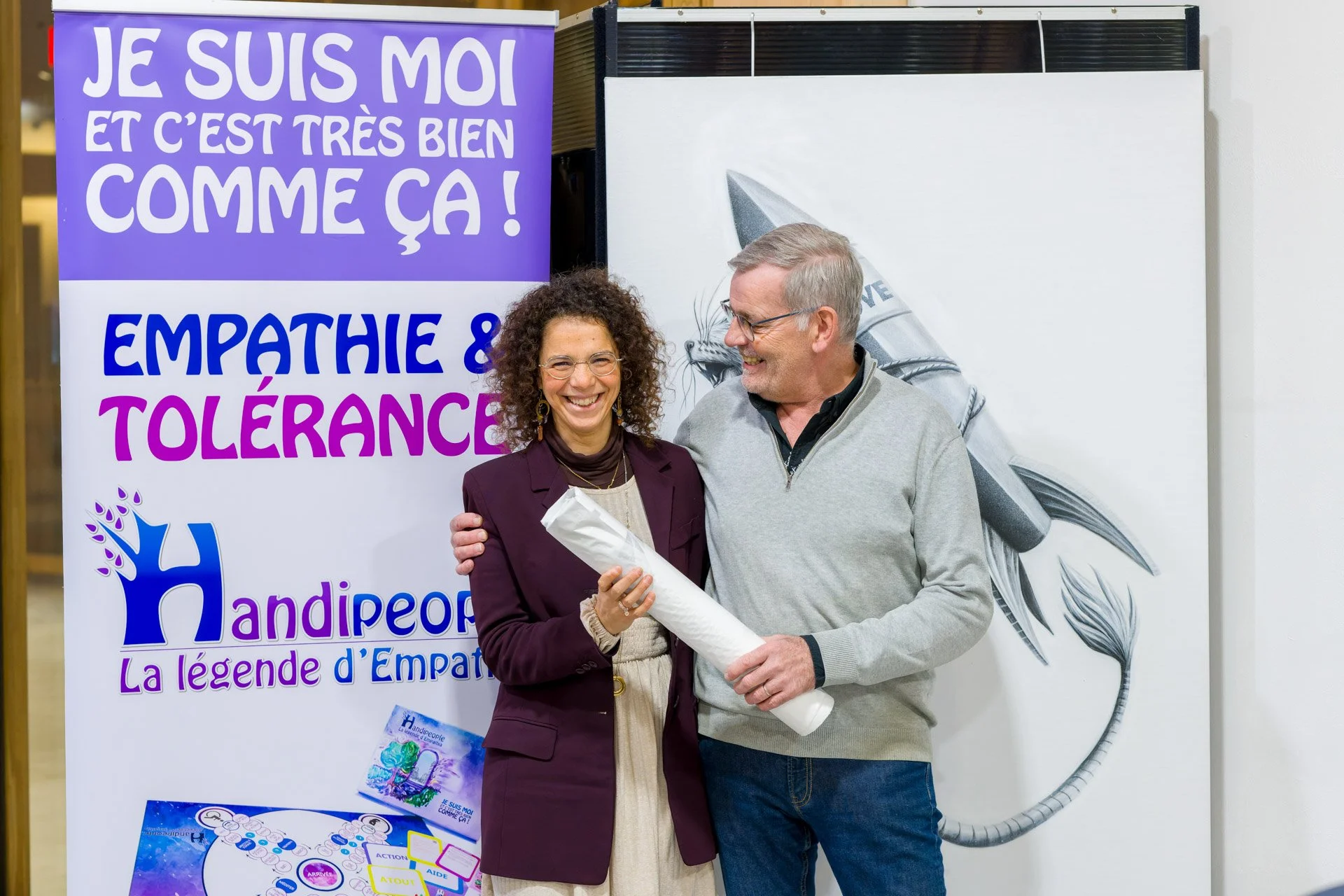 Deux personnes souriant devant une affiche avec le texte "Je suis moi et c'est très bien comme ça ! Empathie & Tolérance". Logo "Handipeople" visible.