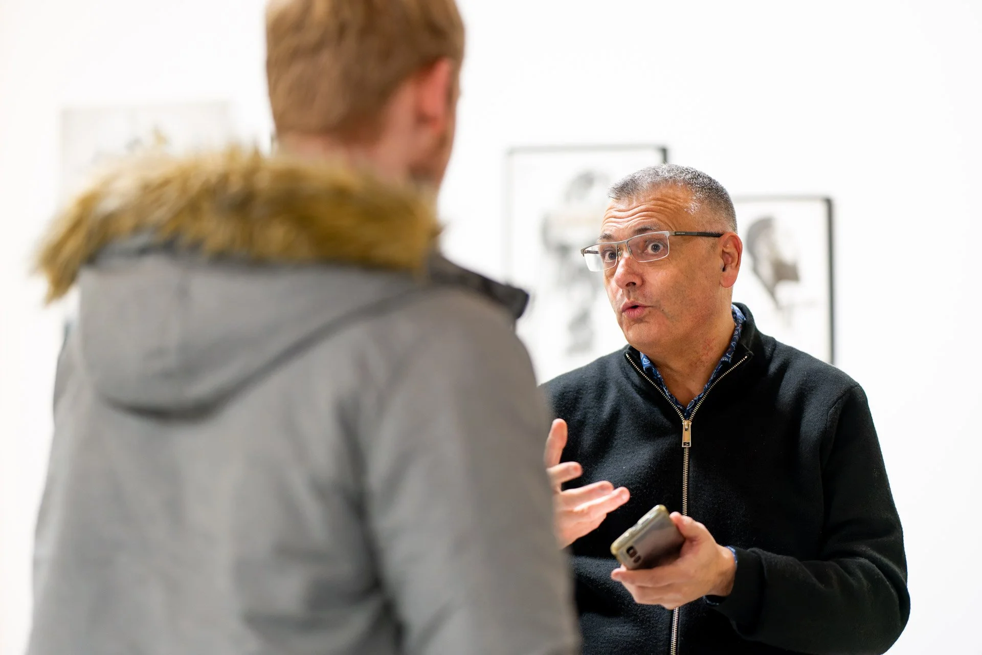 Deux hommes discutant dans une galerie d'art, l'un tenant un smartphone.