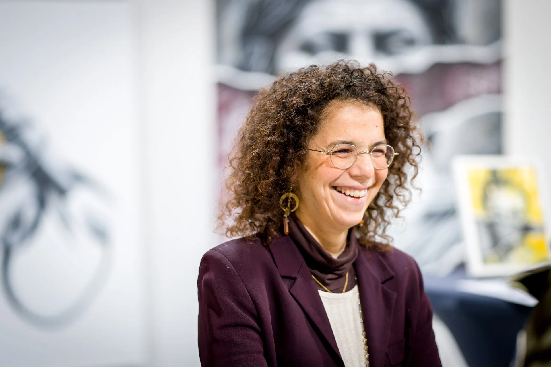 Une femme souriante aux cheveux bouclés, portant des lunettes et un blazer, est debout devant une œuvre d'art floue en arrière-plan.