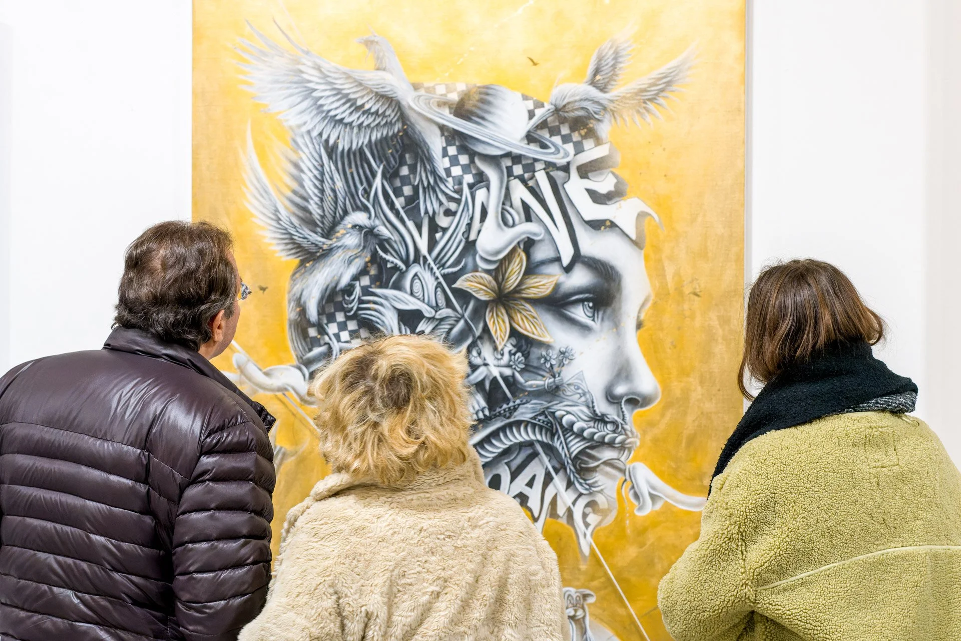 Trois personnes regardent une peinture murale aux motifs complexes avec des éléments naturels et un visage humain stylisé sur un fond doré.