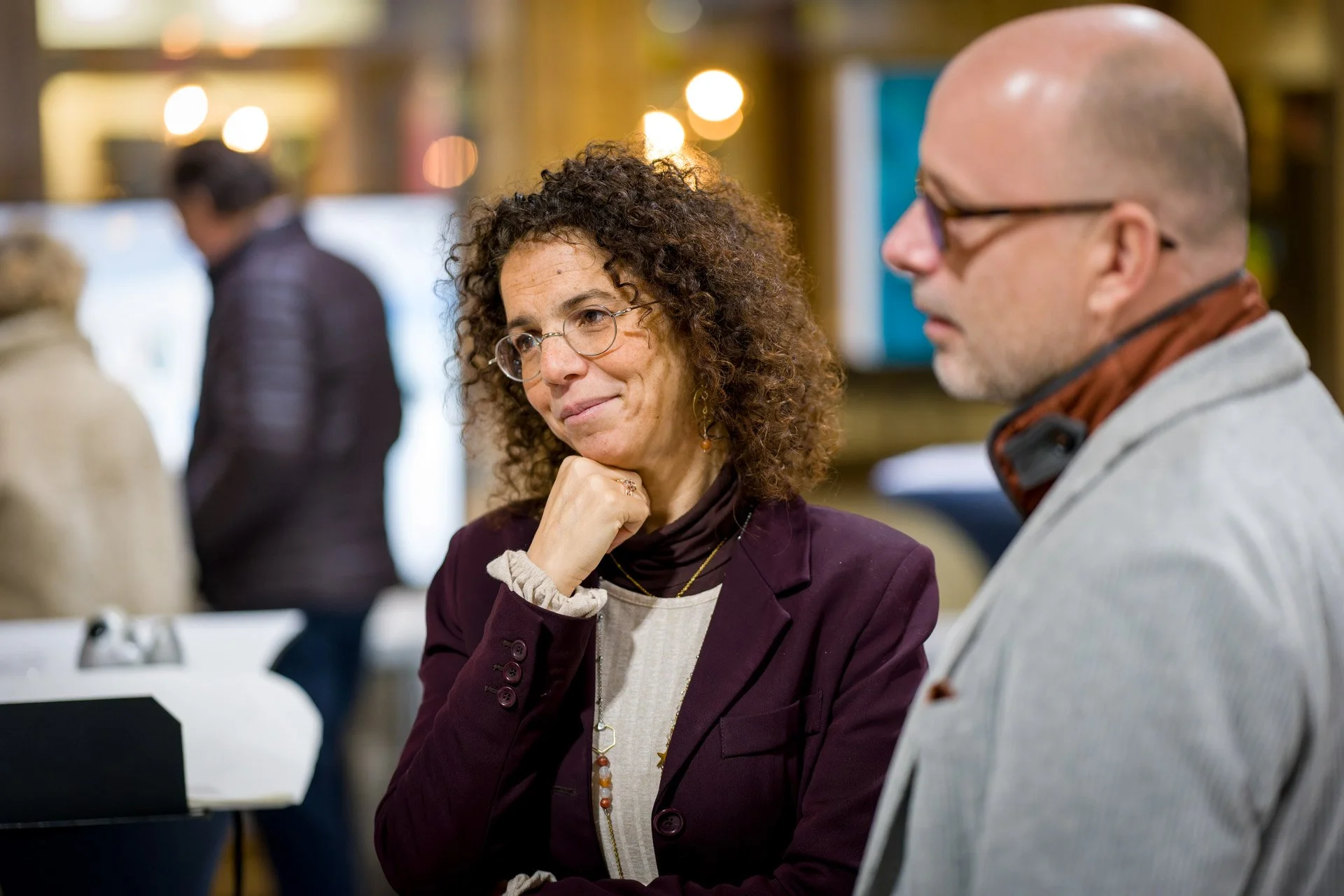 Deux personnes en conversation dans un lieu intérieur, une femme avec des lunettes rondes, cheveux bouclés, portant une veste violette et une chemise beige, semble attentive, un homme plus âgé avec une tête chauve, portant des lunettes et une veste grise, est à ses côtés.