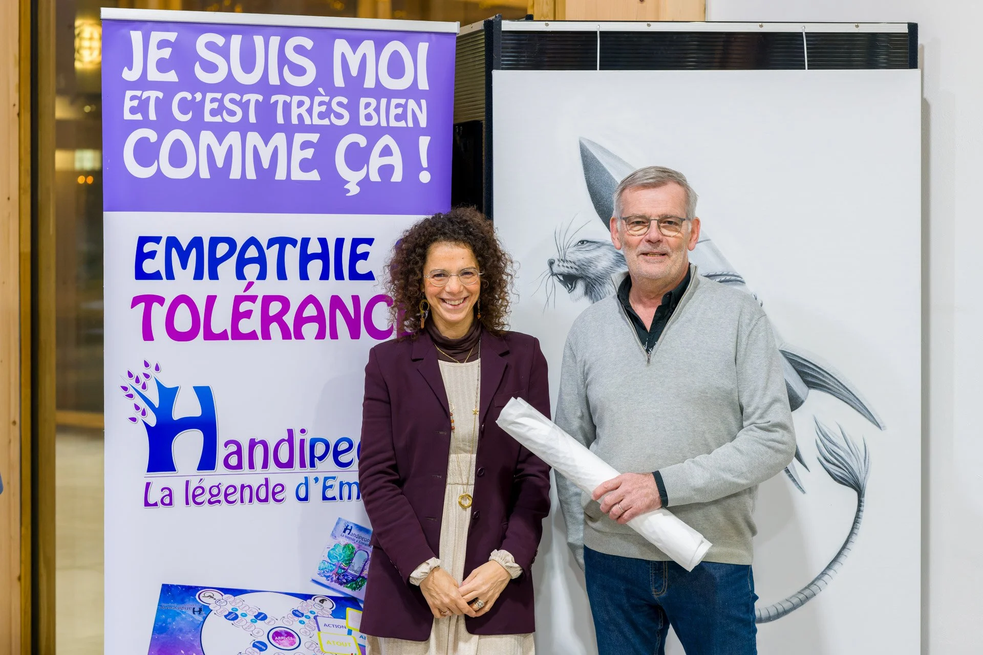Deux personnes souriantes devant une affiche avec le texte "Empathie Tolérance" et "Handipe La légende d'Em". L'homme tient un document roulé.