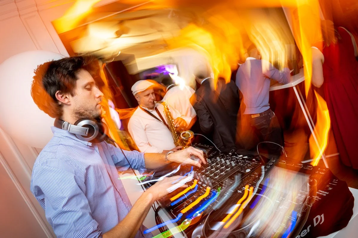 A DJ mixing music at a party or event with guests in the room, a festive atmosphere, coloured lights and people moving around