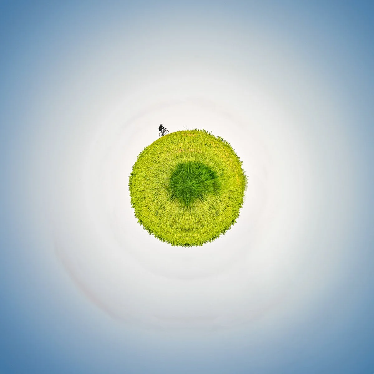Photo professionnelle et artistique d'un cycliste sur une Planète Imaginaire de Paul Marnef, photographe. Cette photo prise aux abors de Louvain-la-Neuve, montre un cysclise qui fait le tour d'une planète. Le centre est vert et le ciel bleu