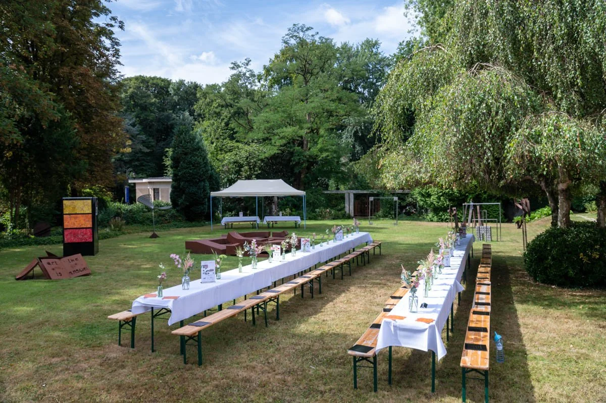 Tables longues avec nappes blanches et bancs en bois installés à l'extérieur dans un jardin verdoyant pour un événement, avec des vases de fleurs et un pavillon en arrière-plan.