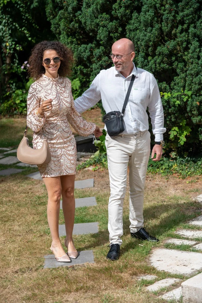 Un homme et une femme marchant dans un jardin, la femme porte une robe beige et des lunettes de soleil, l'homme est en chemise blanche et porte un sac en bandoulière.