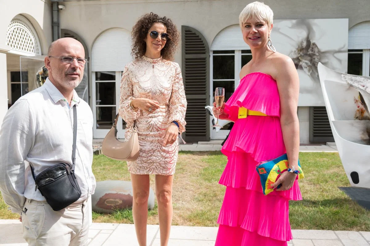 Trois personnes vêtues élégamment à un événement en plein air, une femme portant une robe rose vif tenant un verre de champagne, un homme avec une chemise blanche et une sacoche noire, et une autre femme en robe crème tenant un sac beige. Ils sont de