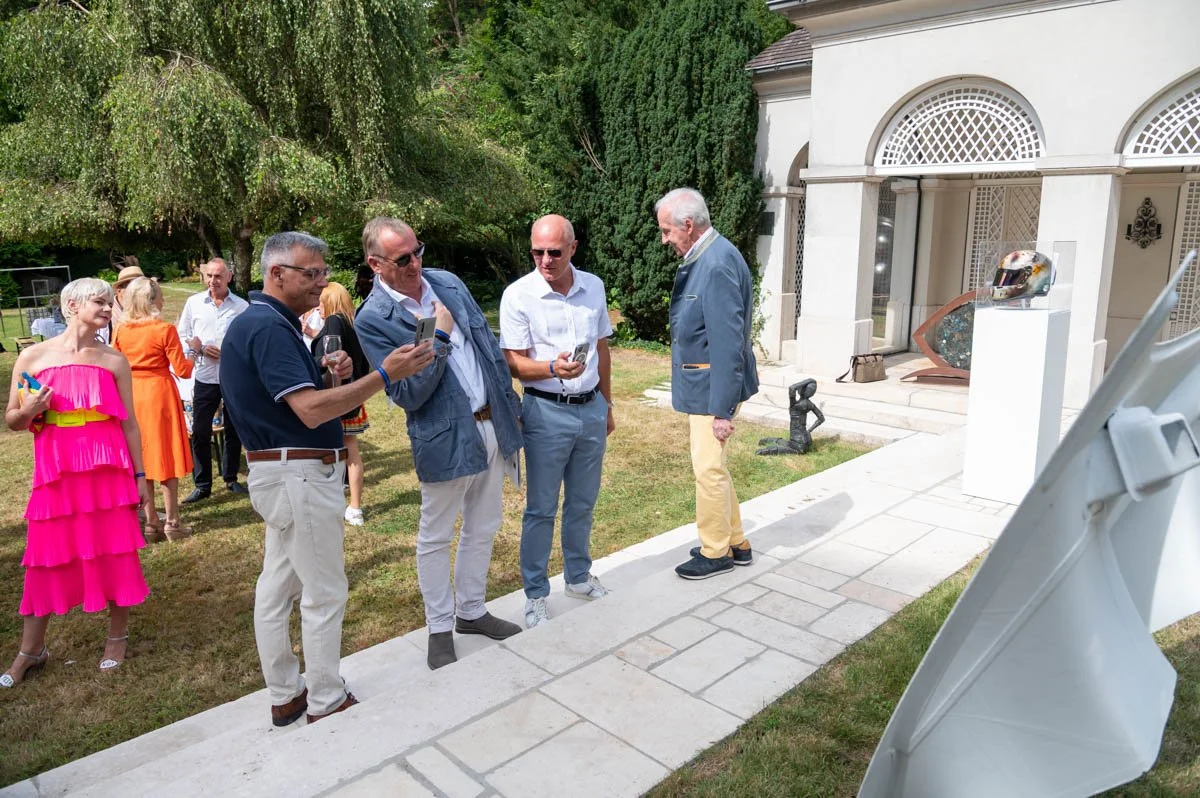 Un groupe de personnes lors d'un événement en plein air. Certaines regardent des téléphones, d'autres discutent. Deux personnes portent des vêtements colorés devant un bâtiment avec des arches. Un casque est exposé sur un piédestal.