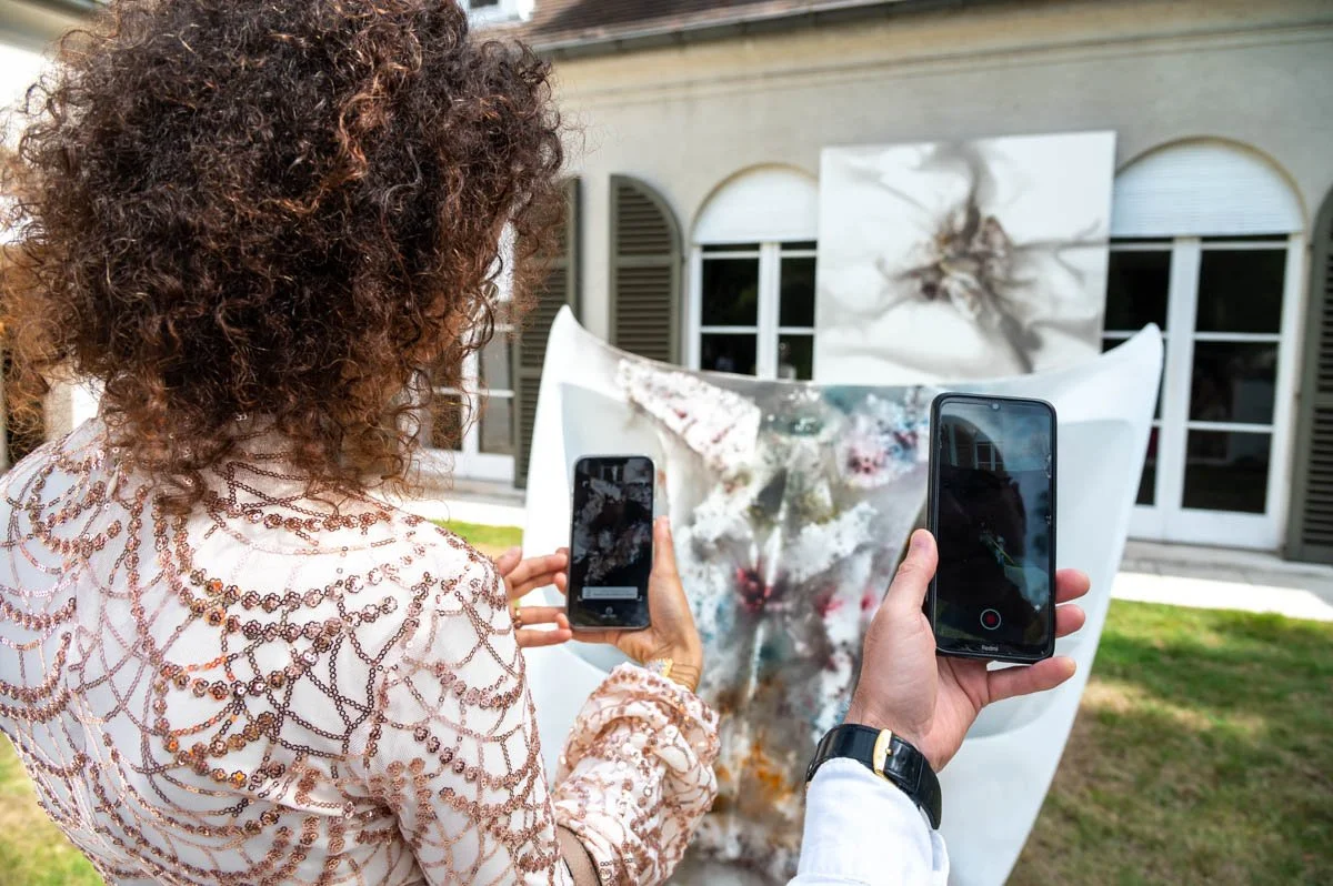 Deux personnes prennent des photos avec des téléphones portables d'une œuvre d'art exposée devant un bâtiment avec des fenêtres. Le chef-d'œuvre est abstrait et coloré.