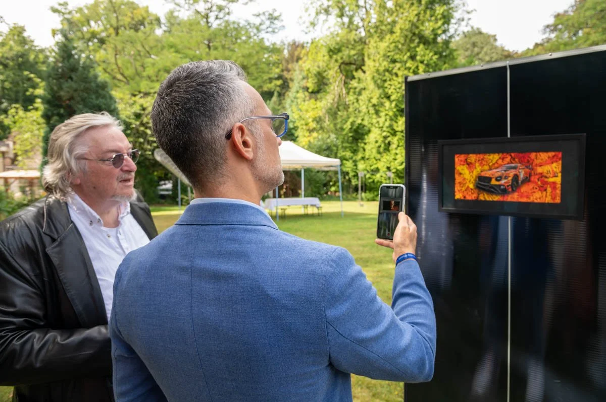 Deux hommes regardent une peinture d'une voiture exposée en extérieur dans un parc verdoyant, l'un d'eux prend une photo de l'œuvre avec un smartphone.
