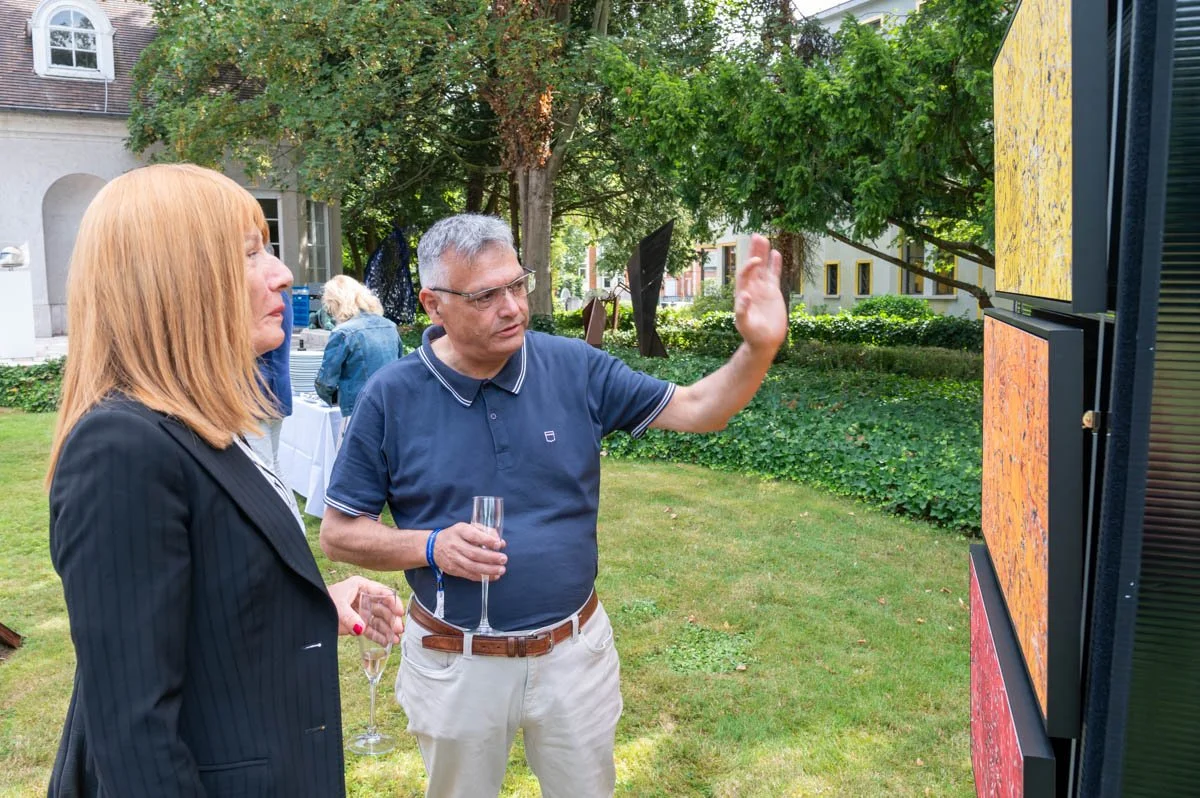 Deux personnes discutant devant une œuvre d'art en plein air, avec des arbres en arrière-plan.